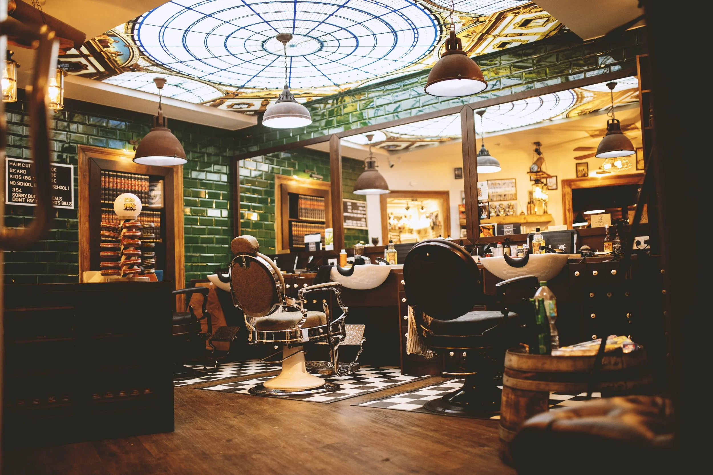 Ein Friseursalon mit klassischen Friseurstühlen, Waschbecken und spiegeln, grünen Fliesenwänden, dekorativen Lampen und einem bemalten Glasdecke.