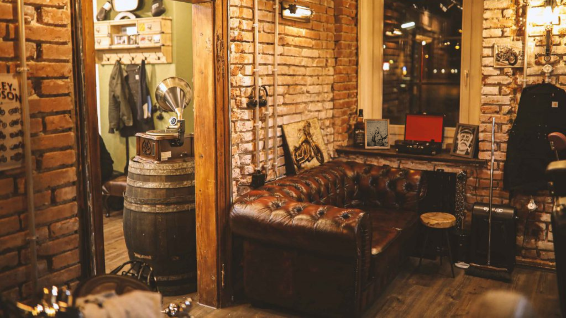 Gemütliche Wohnzimmer-Ecke mit braunem Ledersofa, Holzfußboden, Wand mit sichtbaren Backsteinmauern, Bilder und Dekorationen, Fenster mit Dunkelheit draußen, Vintage- und Musikinstrumente