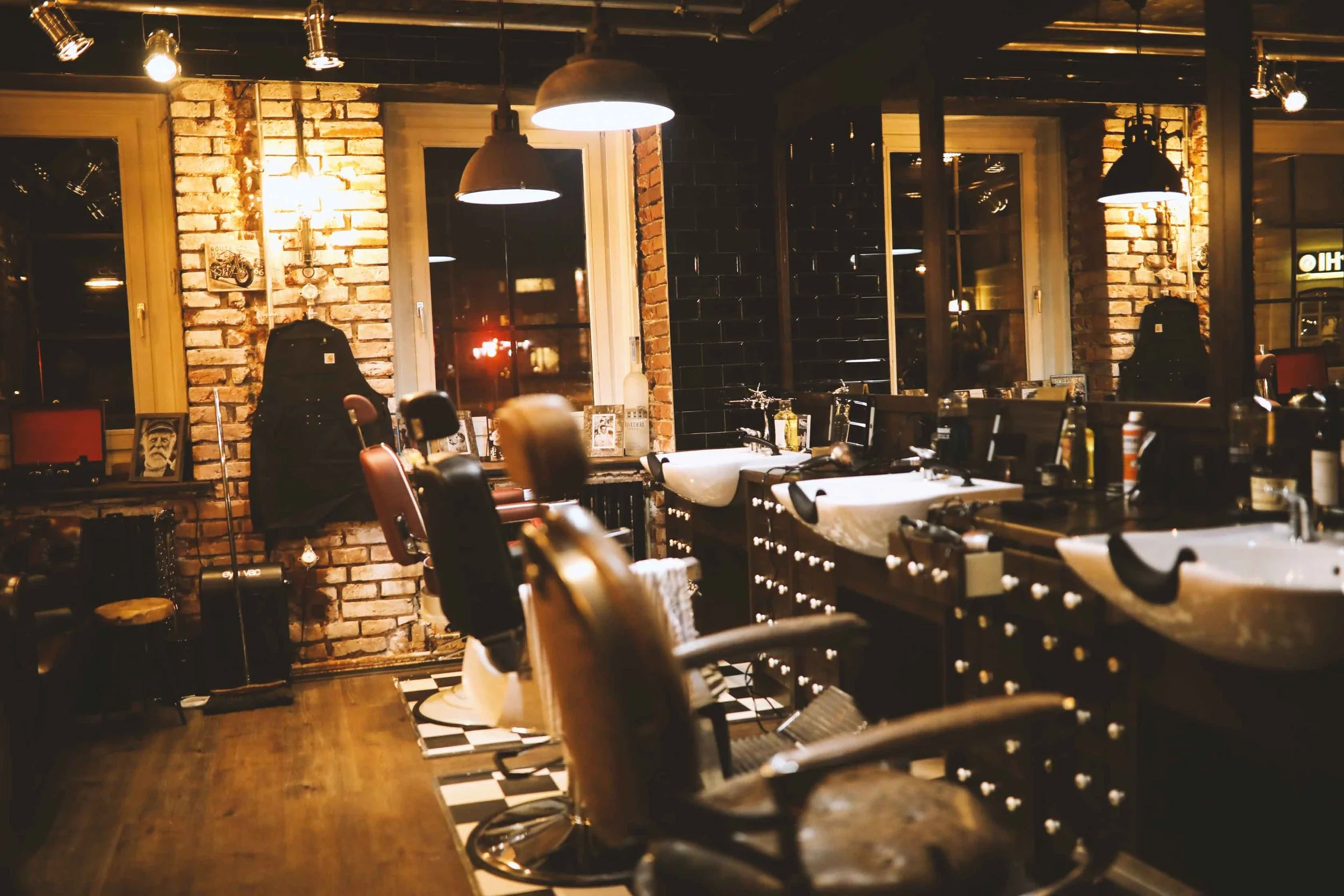 Ein gemütliches Friseurstudio mit beheizten Friseurstühlen, Spiegeln und Waschbecken, eingerahmt von roten Backsteinwänden und großen Fenstern, beleuchtet durch hängende Lampen.