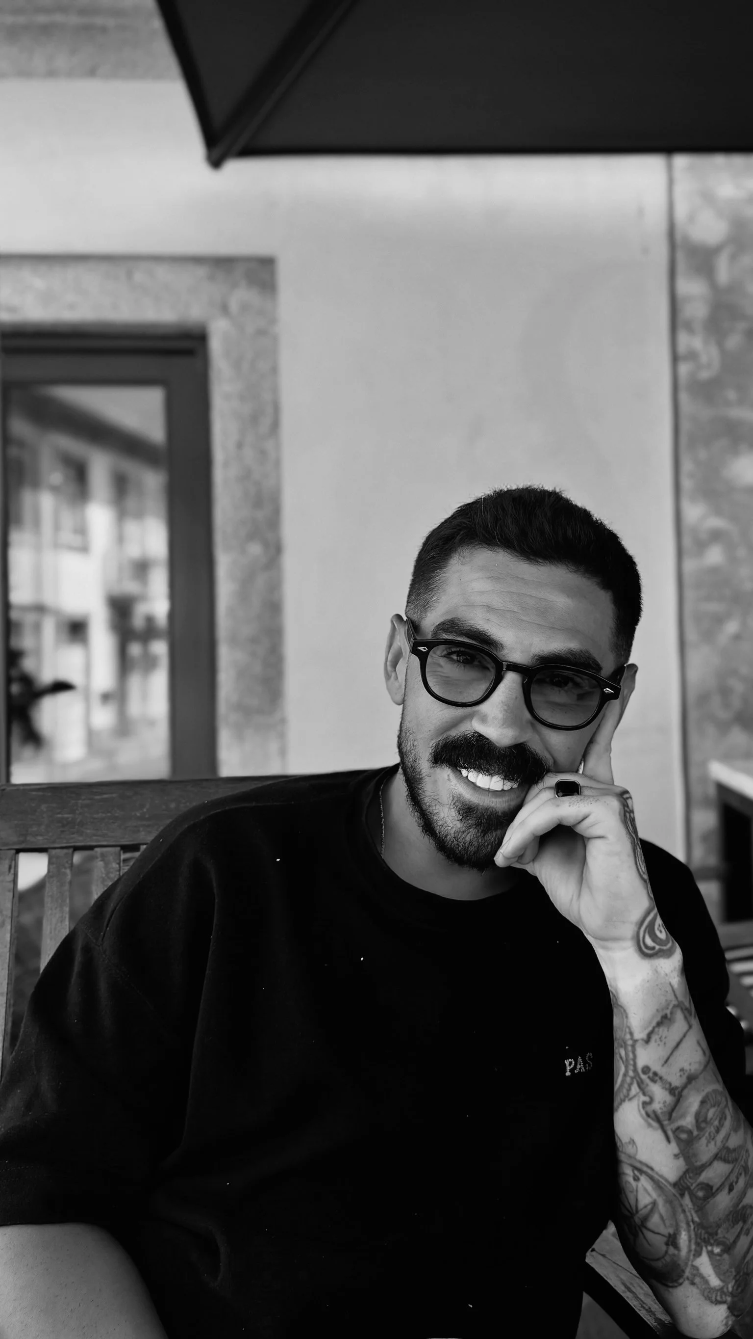 Schwarz-weiß-Foto eines lächelnden Mannes mit Brille, Tattoos am Arm, sitzend auf einer Bank, im Hintergrund Fenster und Außenansicht