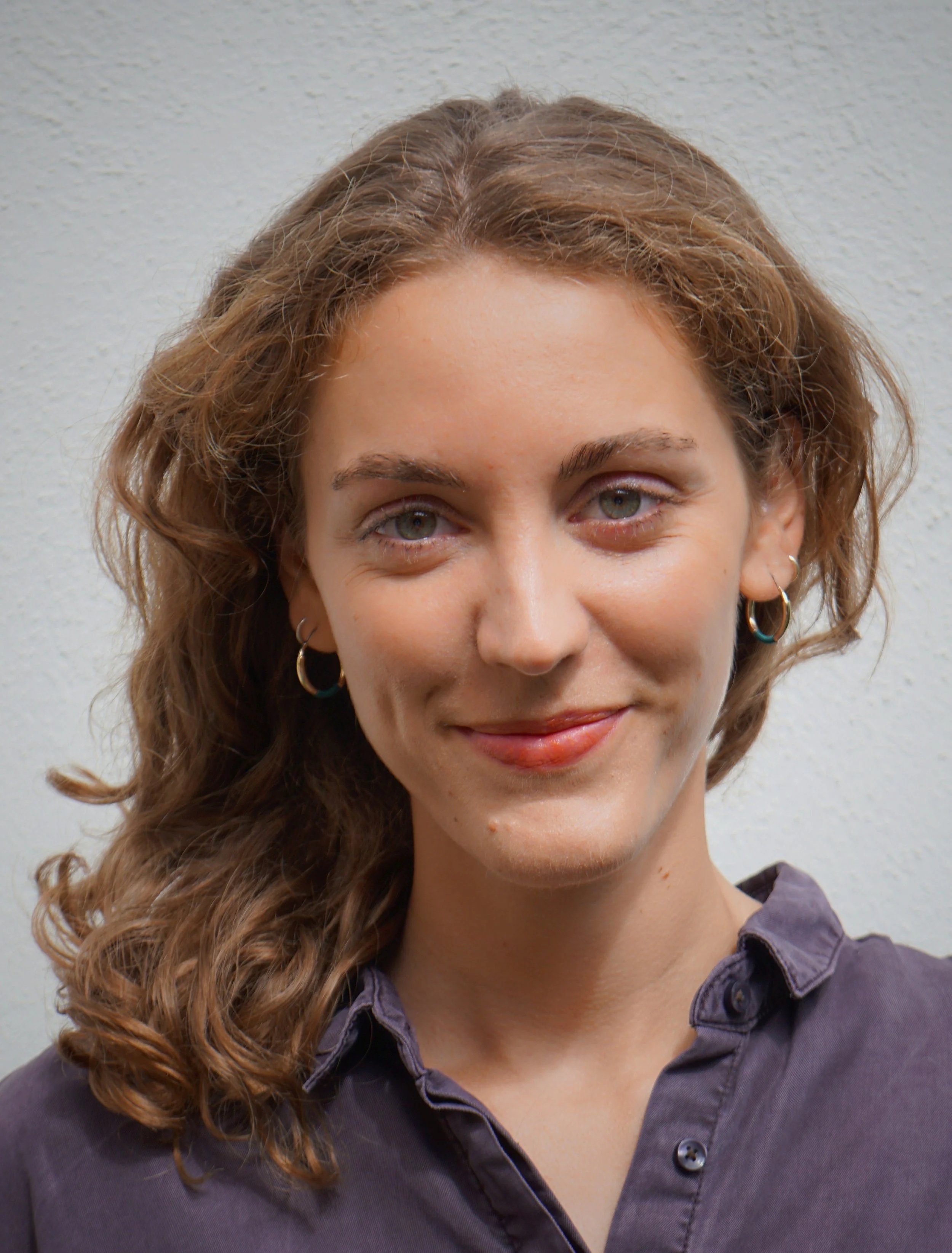 Close-up of a young woman with curly light brown hair, wearing small hoop earrings and a dark button-up shirt, smiling at the camera against a light gray wall.