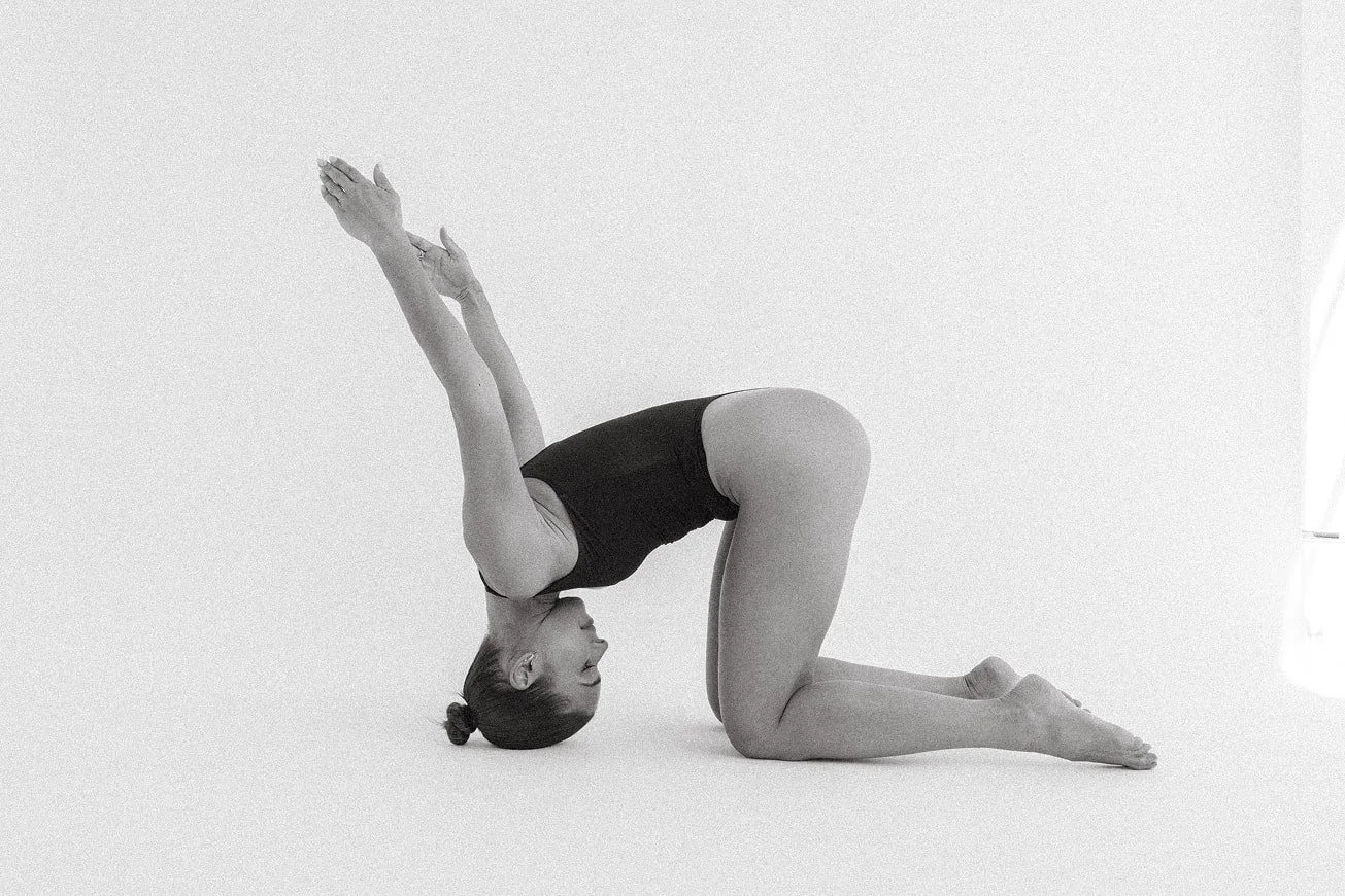A woman practicing yoga, positioned with her hands and forehead on the ground, hips raised, and knees bent, in an indoor studio with a white background.