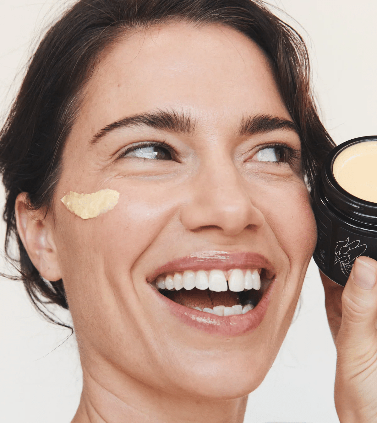 A smiling woman with cream on her cheek holding an open jar of cream near her face.