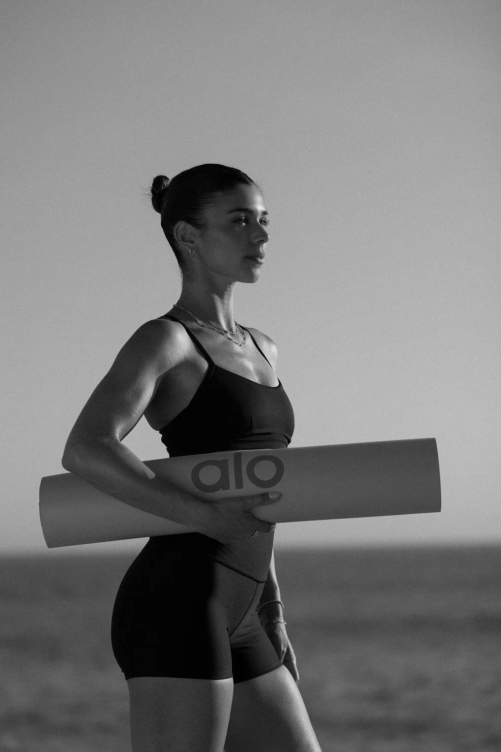 A woman in athletic wear holding a yoga mat on the beach, with the ocean and a clear sky in the background, captured in black and white.