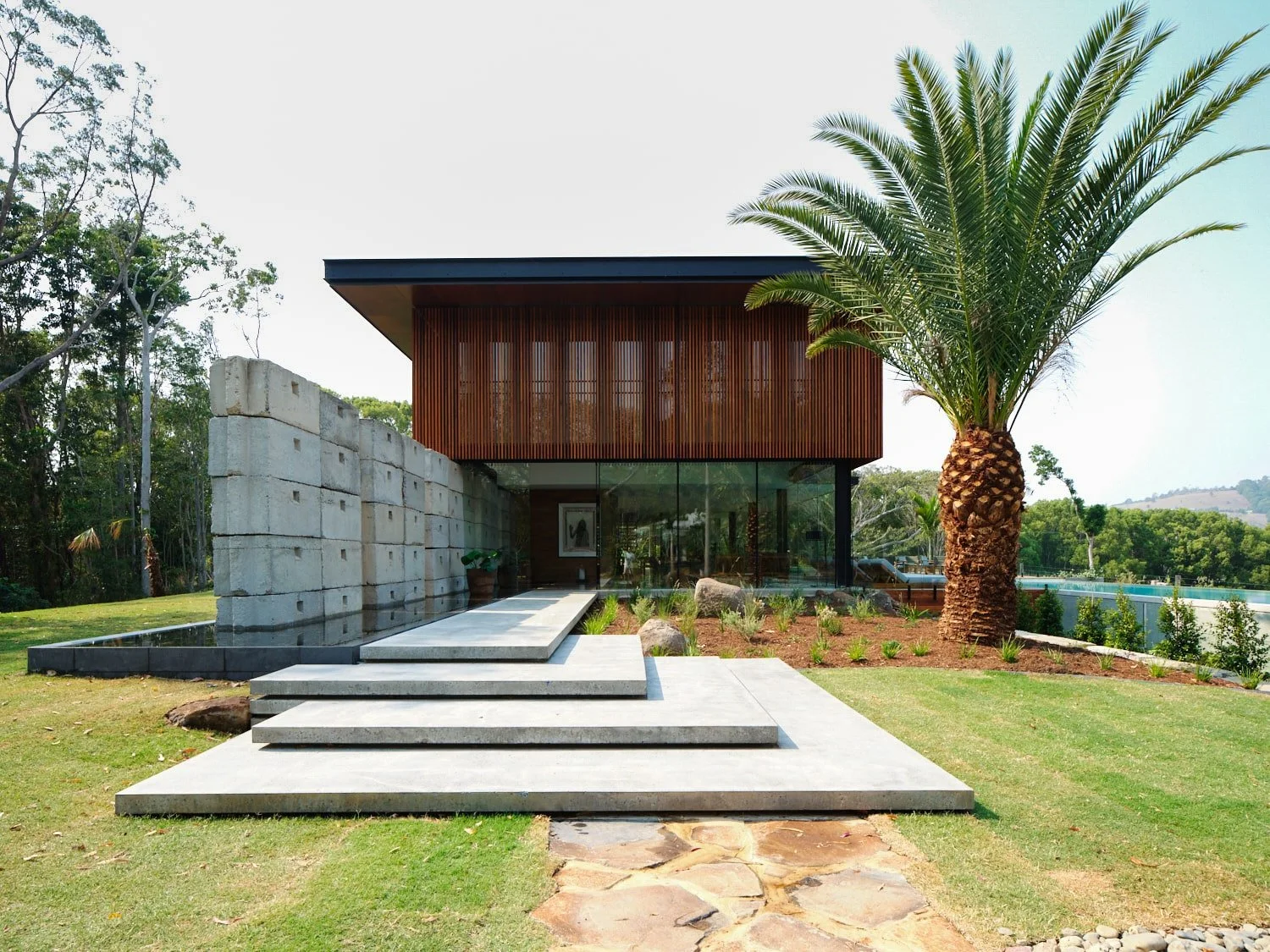 Modern house with glass front, wooden upper walls, concrete stairs, lush green lawn, palm tree, and surrounding natural landscape.