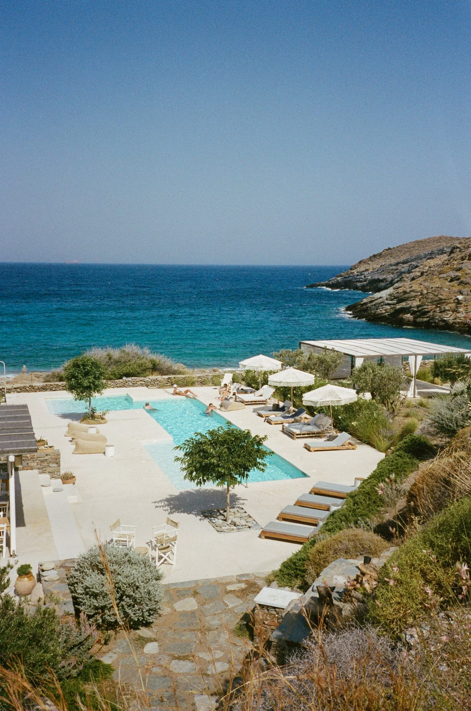 A scenic view of a coastal resort with a swimming pool, lounge chairs, umbrellas, and a rocky shoreline overlooking the ocean on a clear sunny day.