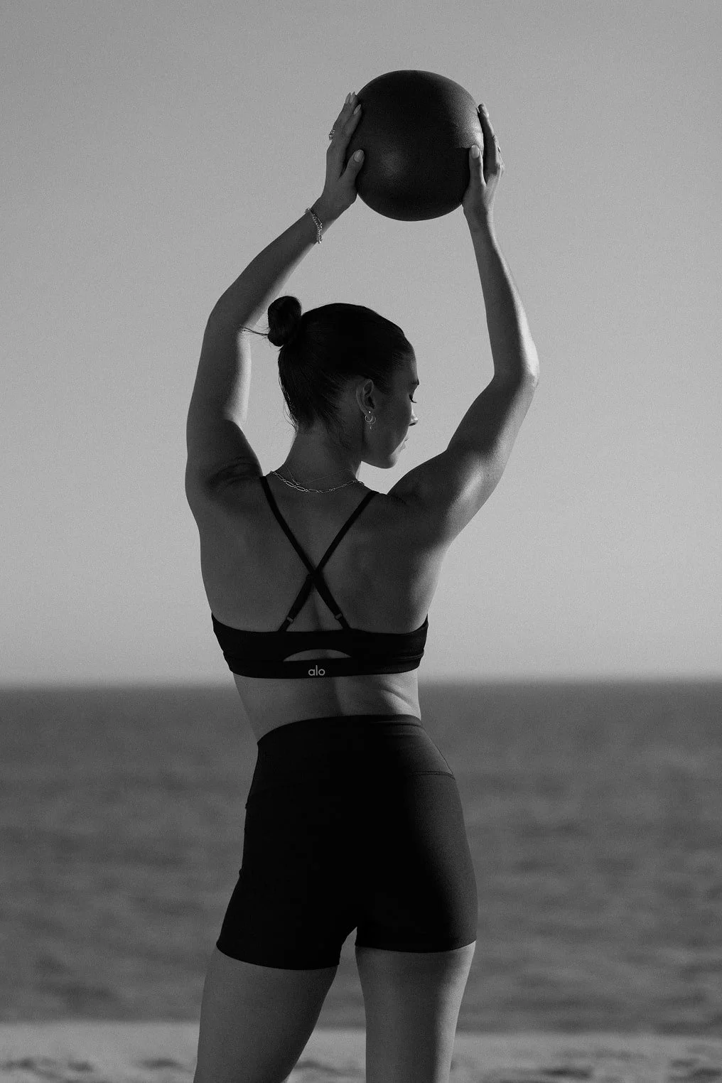 A woman standing outdoors near water, holding a ball overhead with both hands, dressed in workout attire.