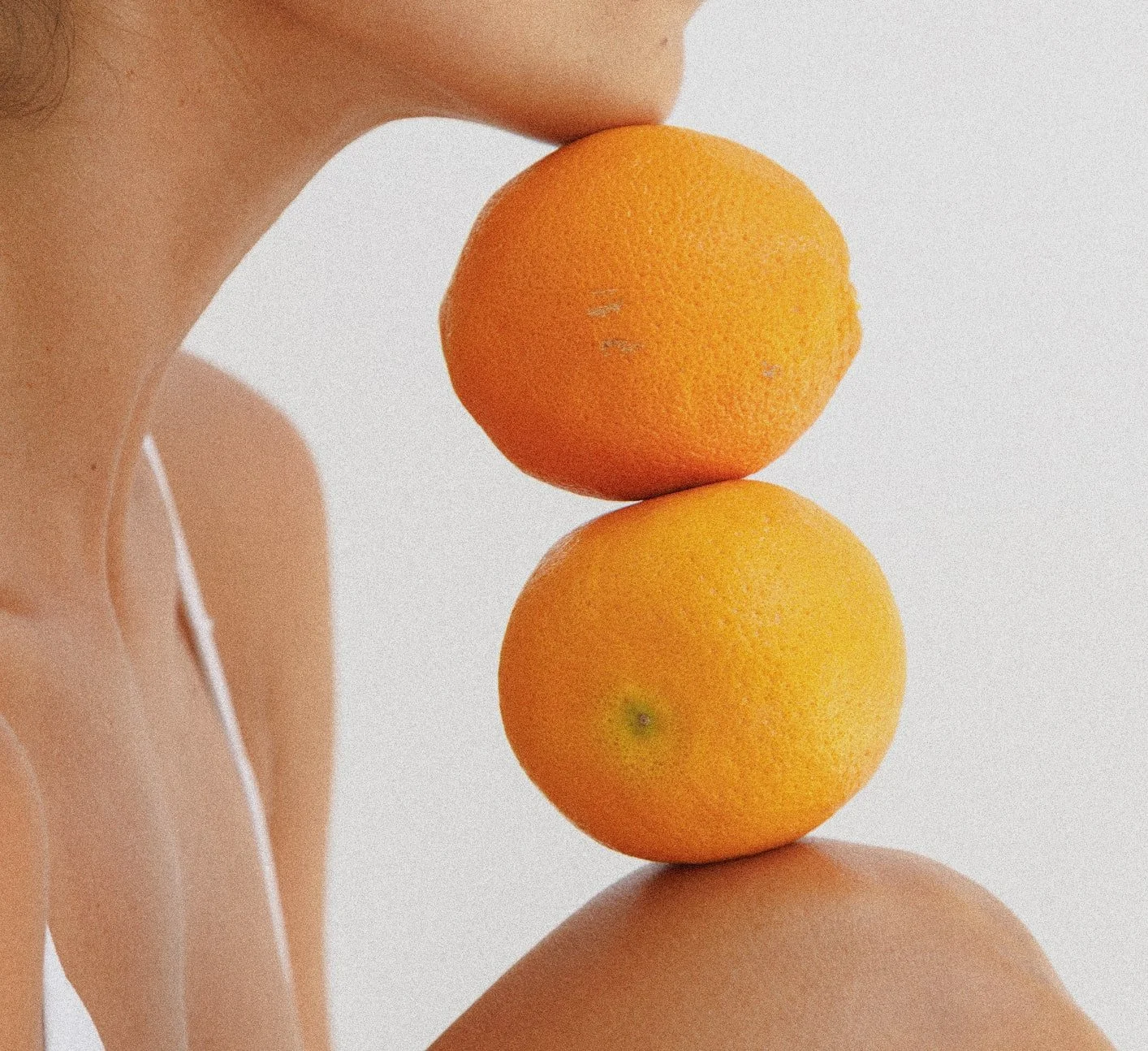 A person balancing three oranges one on top of the other, with their face not visible, wearing earphones, and the background is plain white.