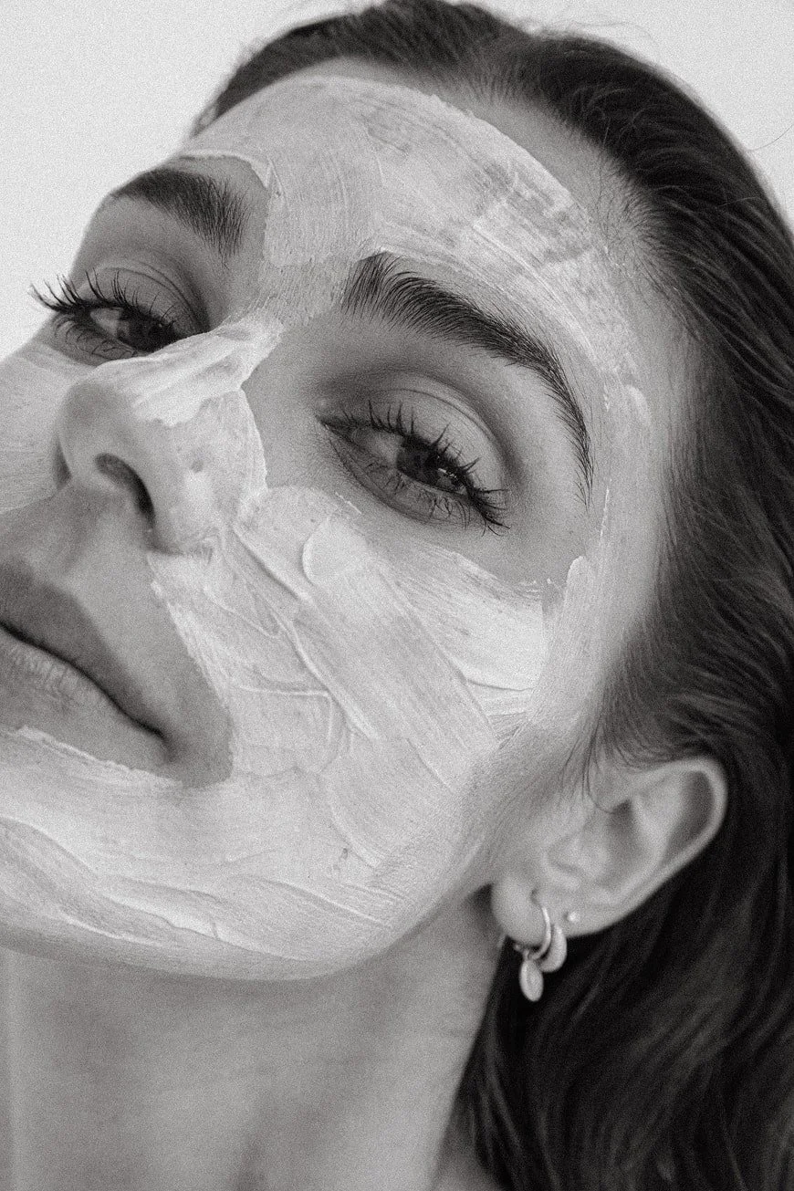A close-up black and white photo of a woman with a face mask applied to her face, her eyes partially closed, and her hair swept back.