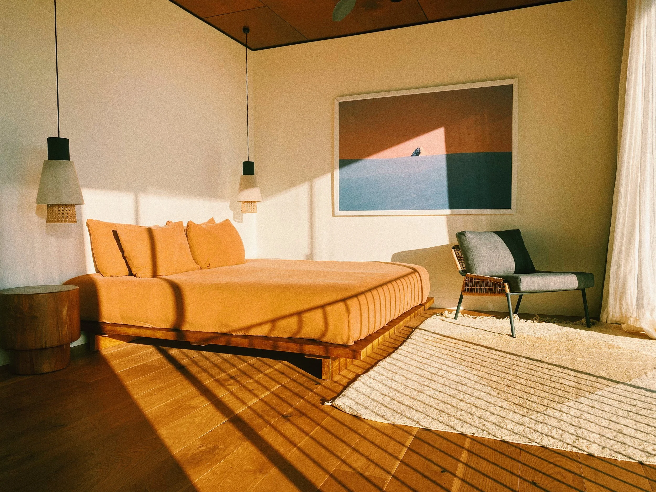 A bedroom with a bed dressed in peach bedding, a small wooden side table, a gray armchair, a large framed artwork on the wall, and curtains drawn to the side, all illuminated by warm natural sunlight.