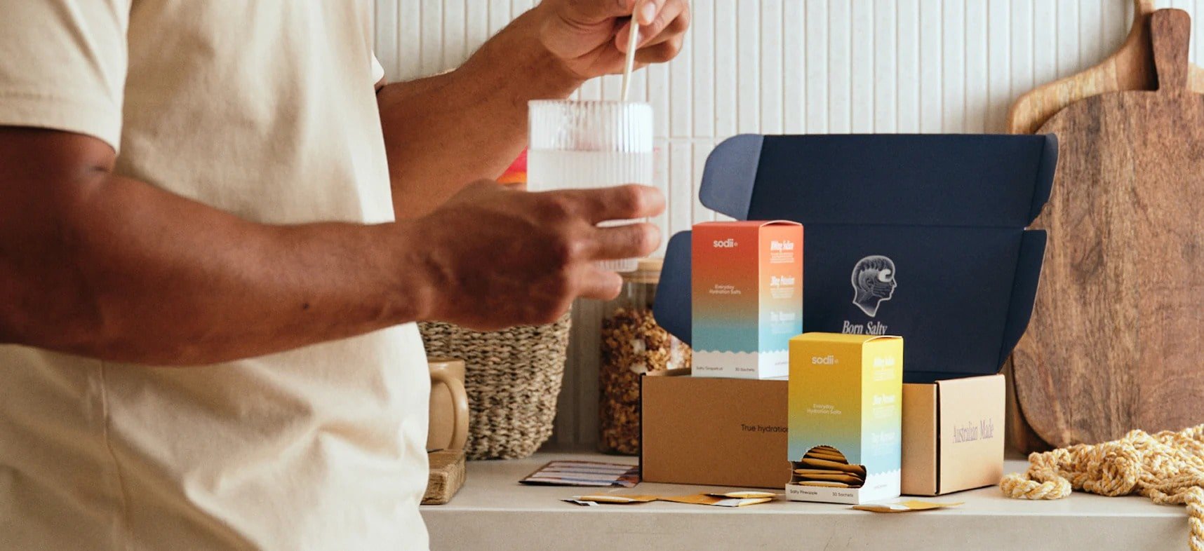 A person holding a glass of a clear beverage with a straw, standing next to a display of colorful boxes labeled 'Soda' and 'True Hydration' on a countertop, with a navy blue bag with a logo and a cutting board in the background.