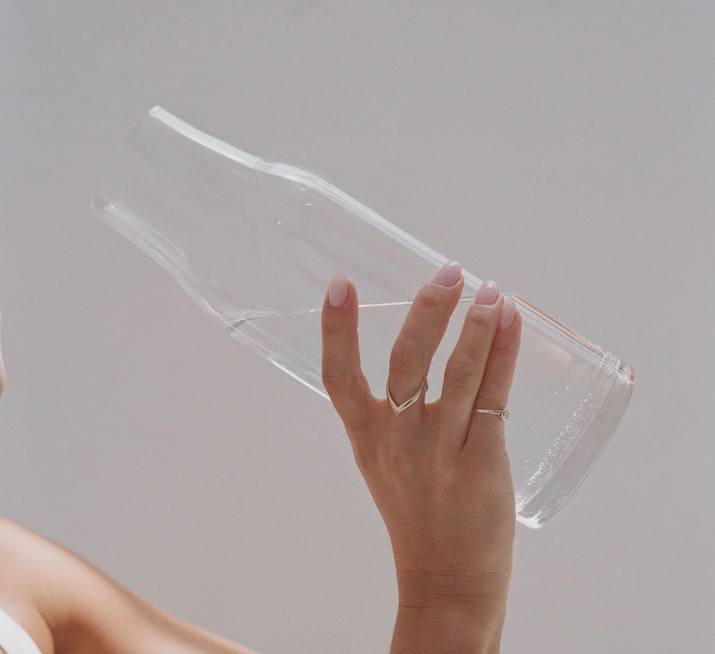 A woman holding a clear, empty glass bottle with her hand, showing a silver ring on her finger, against a plain gray background.