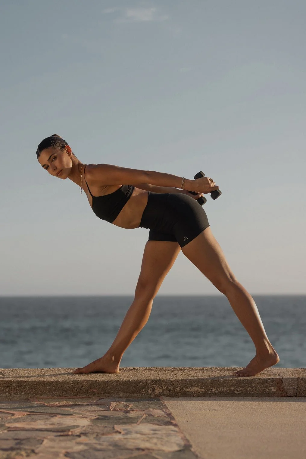 A woman in black workout clothing holding dumbbells backwards, performing a stretching or exercise pose outdoors near the ocean during daytime.