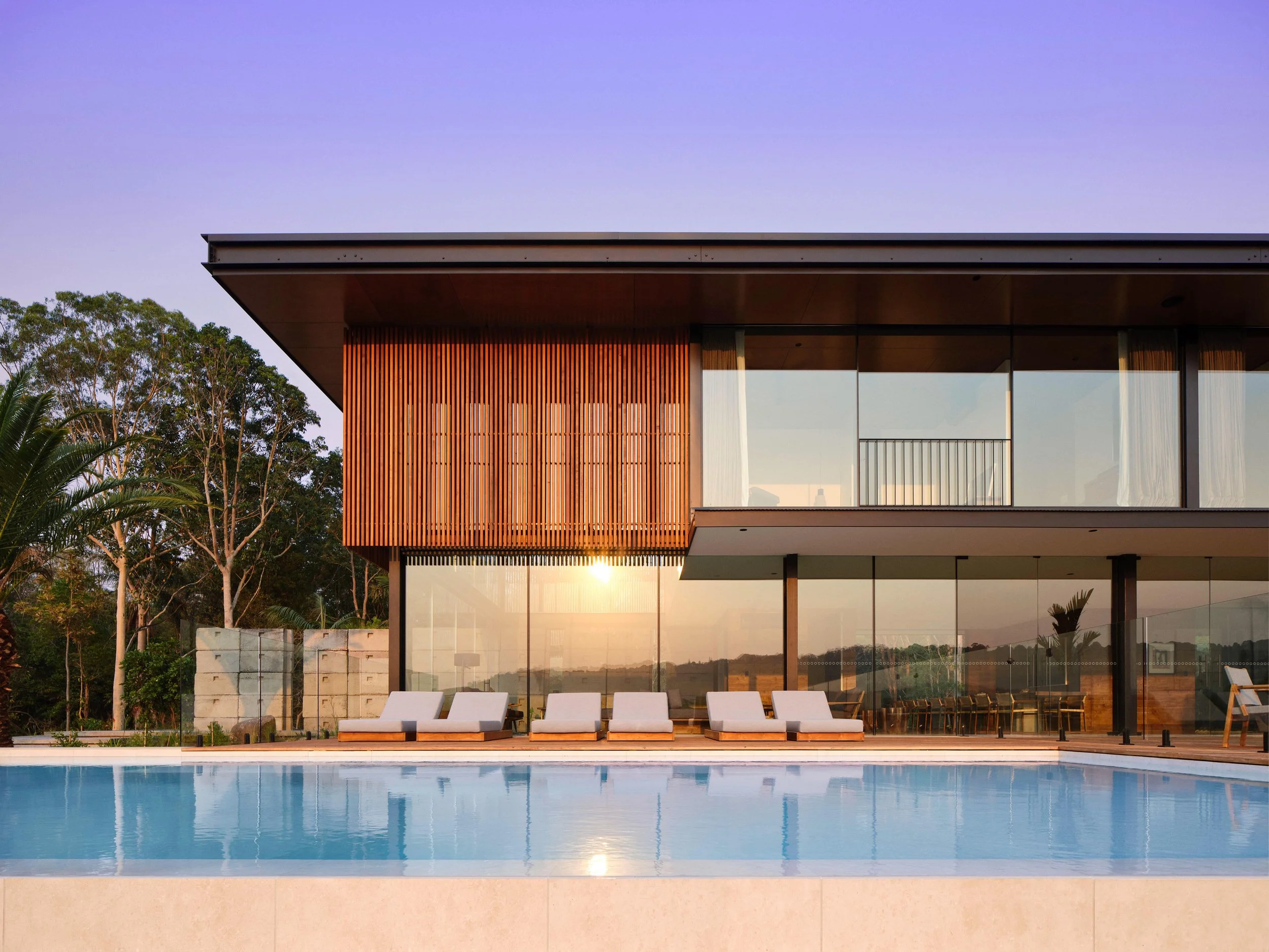 Modern house with large glass windows, a swimming pool with white lounge chairs, and trees in the background during sunset.