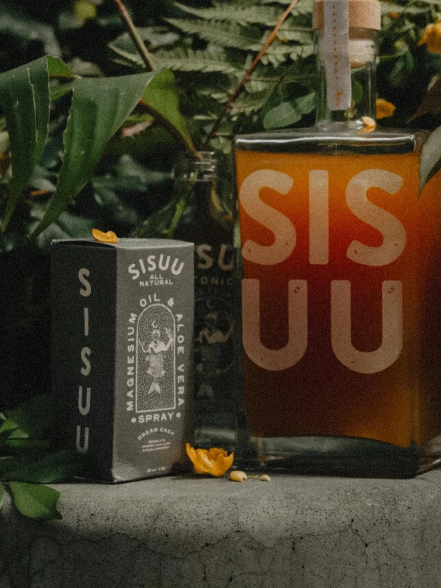 A black carton of Sisuu magnesium oil spray and a glass of orange juice with the words 'SISU' on it, placed on a concrete surface with green foliage in the background.