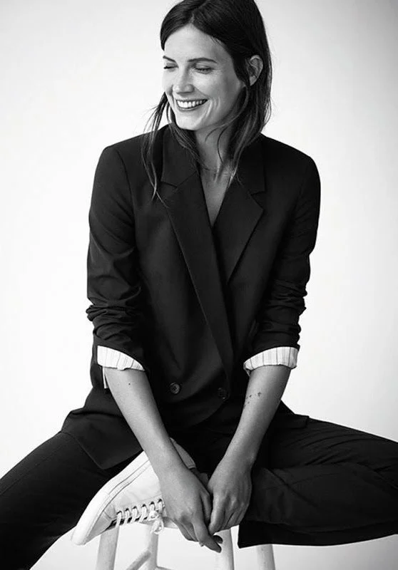 A woman sitting cross-legged on a stool, smiling and looking to her right, wearing a black blazer with rolled-up sleeves and white sneakers.