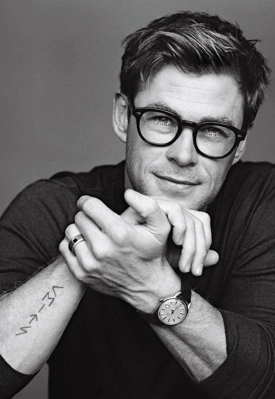 Black-and-white portrait of a young man with glasses, wearing a dark top, with a watch, ring, and tattoo visible, posing with his arms crossed in front of his face.