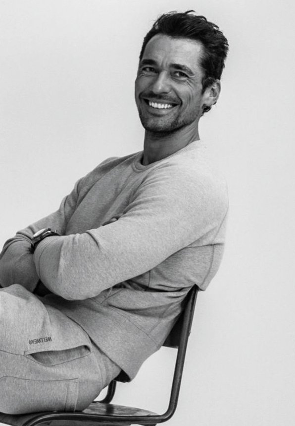 A smiling man with dark hair and a beard, wearing a light-colored long-sleeve shirt, sitting on a chair against a plain white background.
