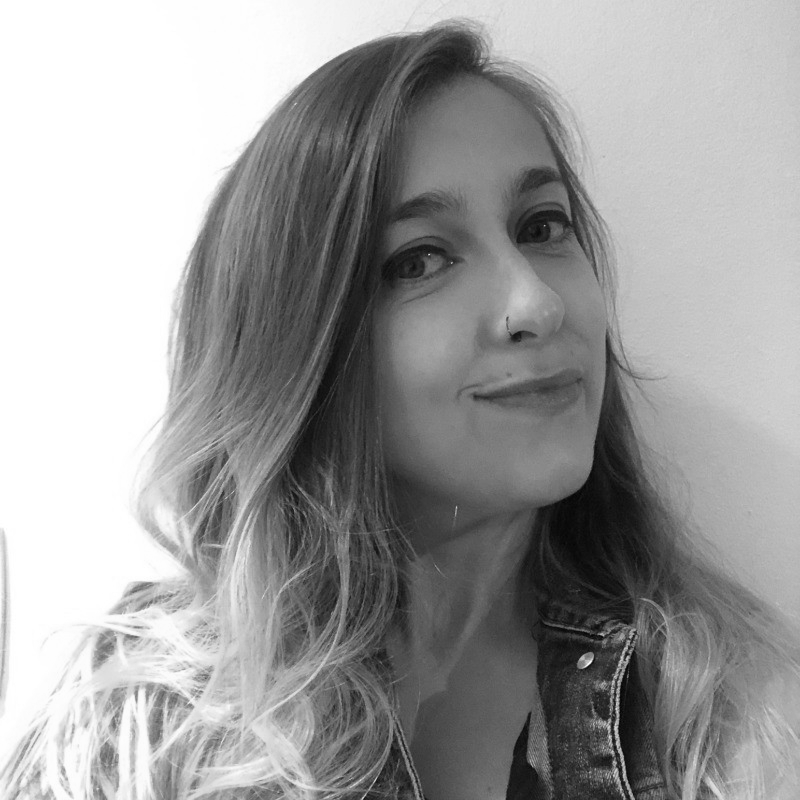 Black and white photo of a young woman with long wavy hair, smiling slightly, wearing a denim jacket and a nose ring.