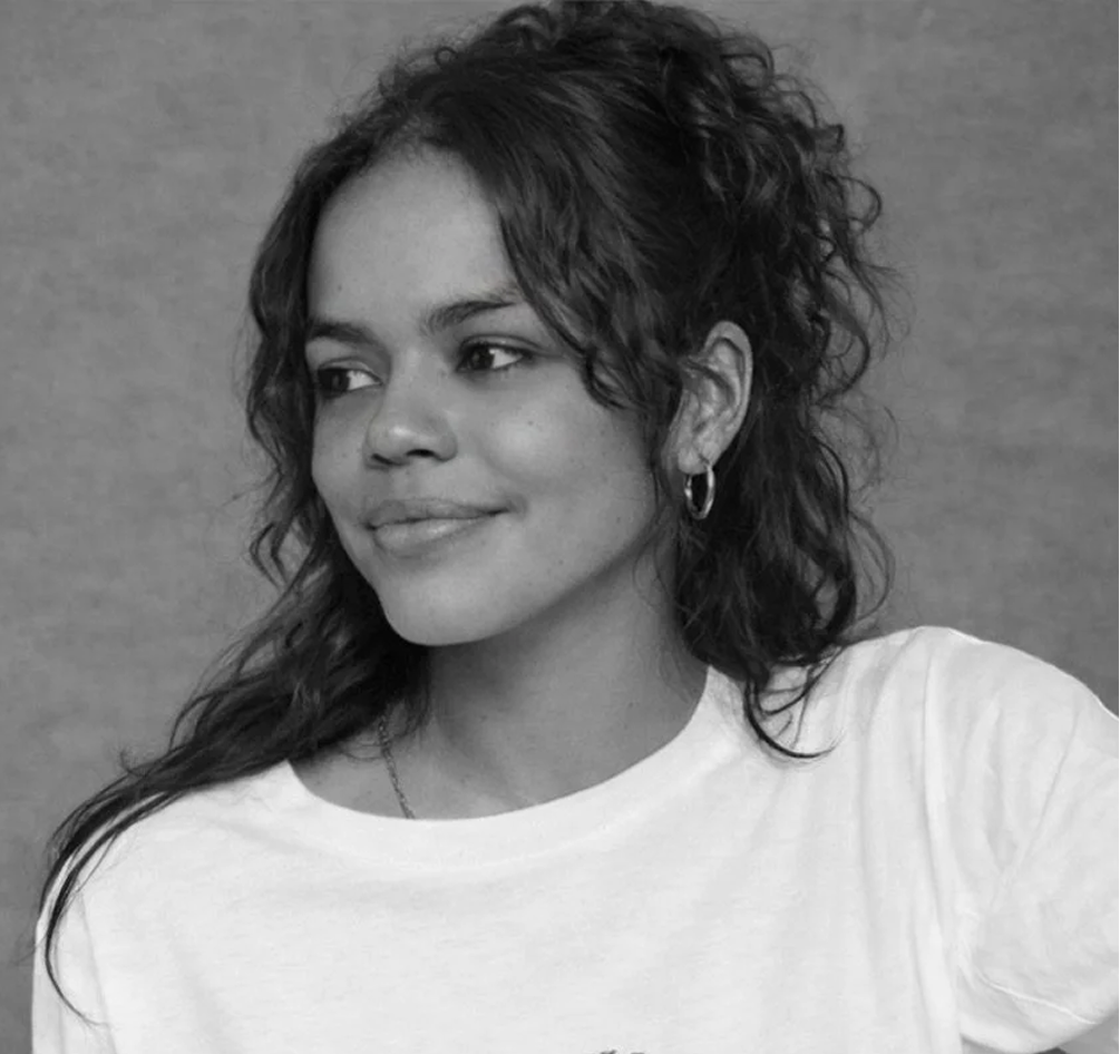 A black-and-white photograph of a woman with curly hair, wearing a white shirt and hoop earrings, looking to her left with a slight smile.