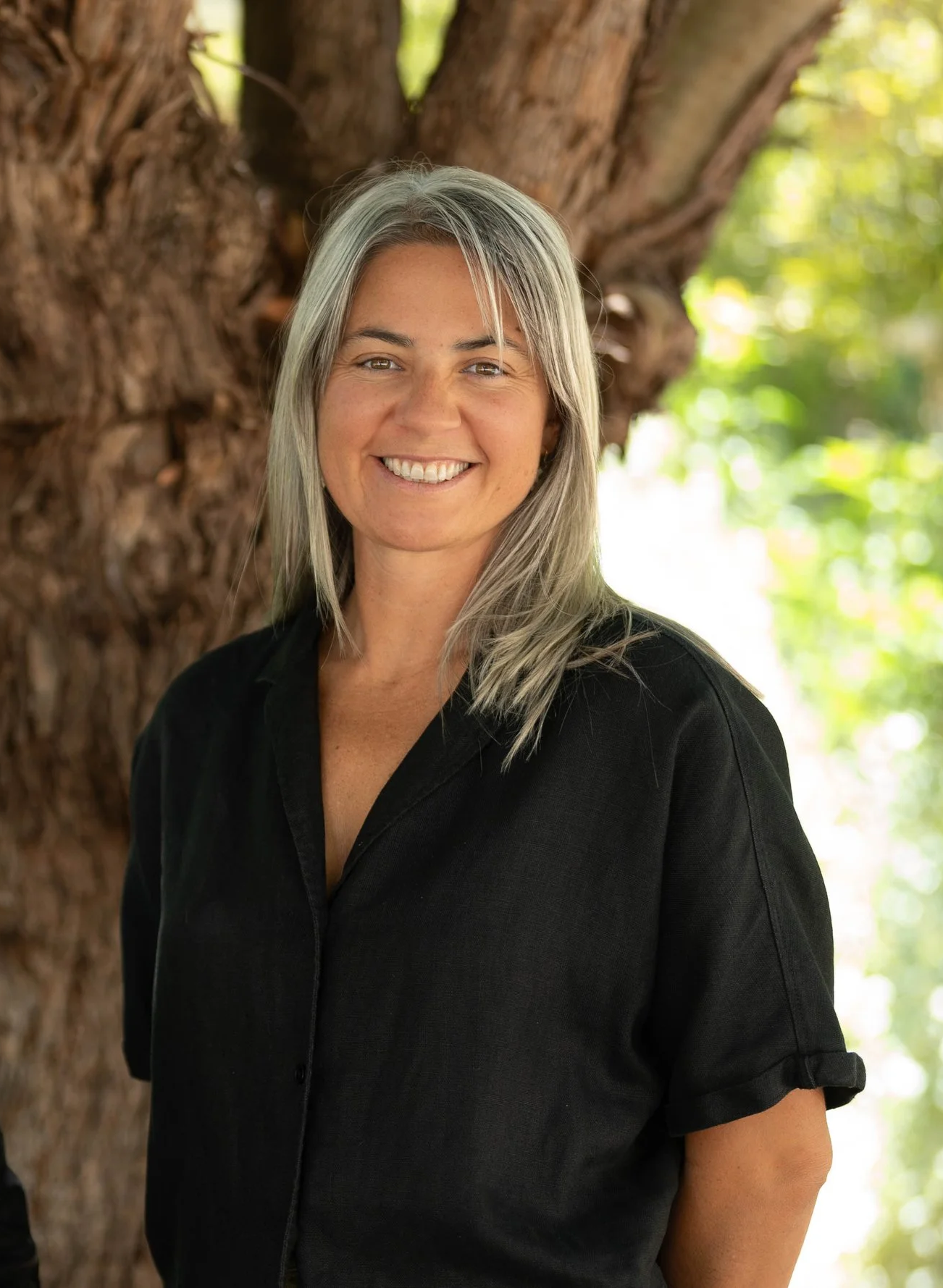 A smiling woman with gray hair wearing a black shirt standing outdoors in front of a large tree with green leaves.