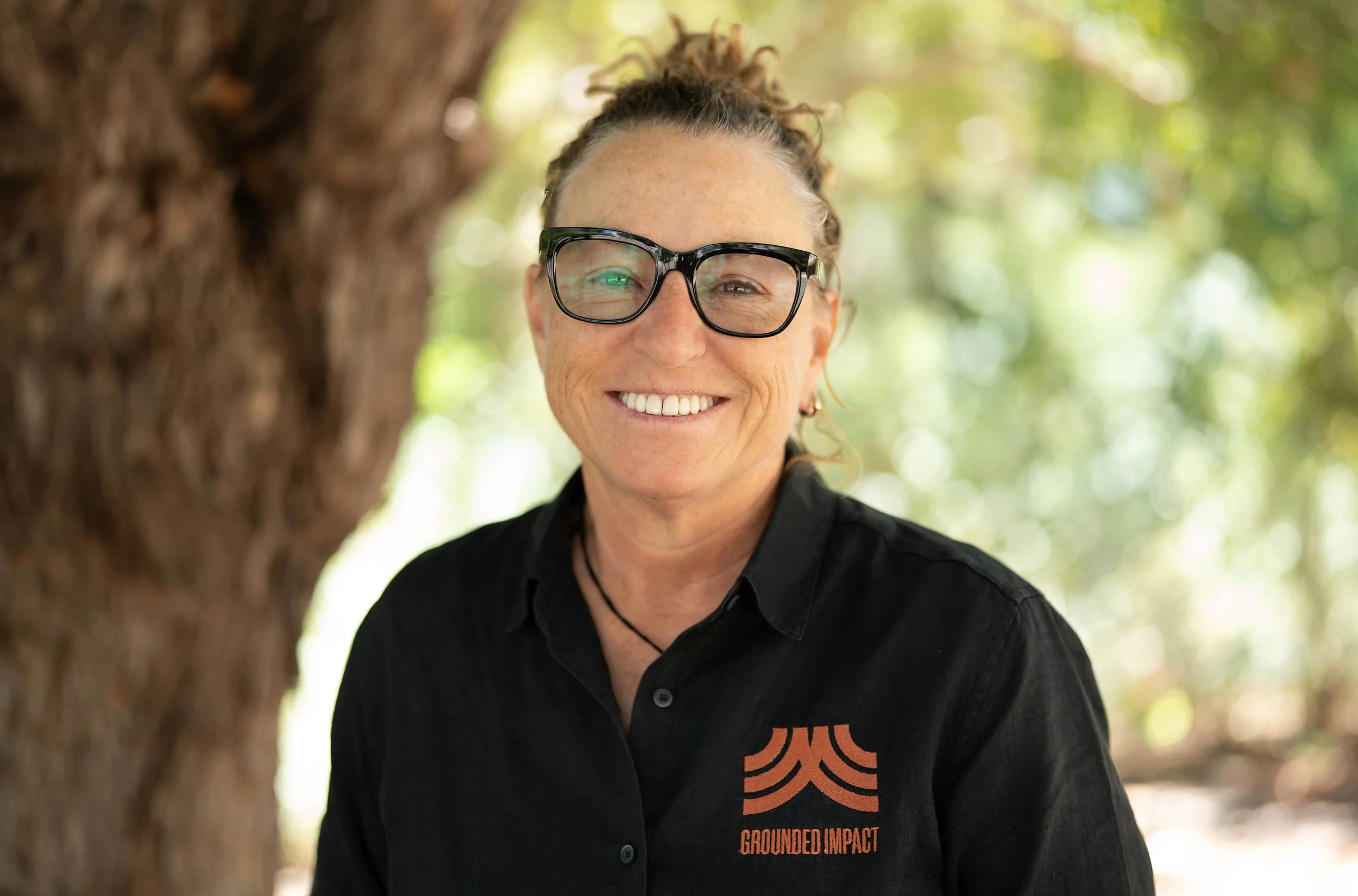 A woman with curly hair tied up, wearing glasses and a black shirt with an orange logo that says 'Grounded Impact', smiling outdoors with a blurred background of green trees and sunlight.
