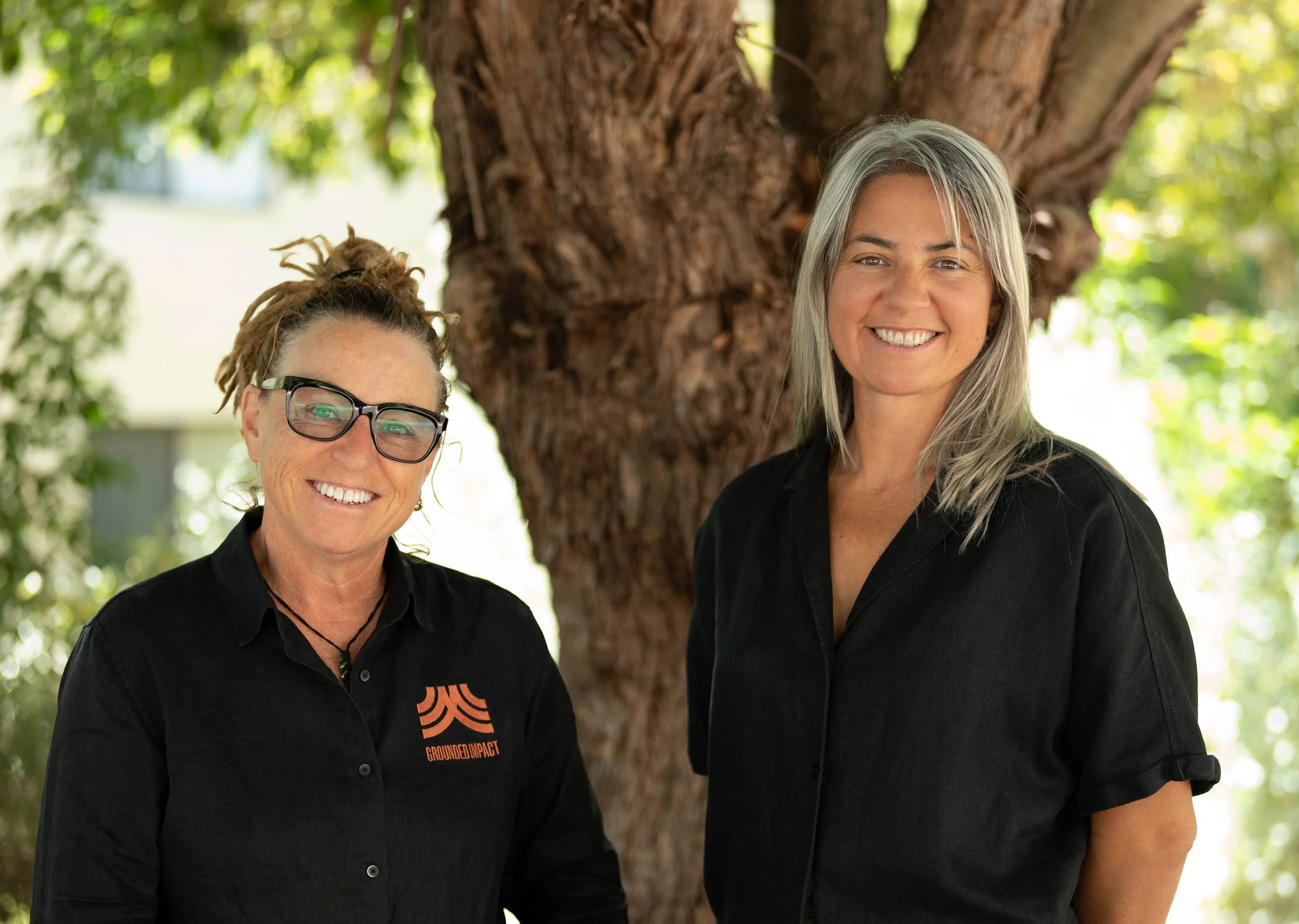 Two women standing outdoors next to a tree with green leaves in the background. They are smiling and wearing black shirts, one with glasses and a bun hairstyle, the other with long gray hair.