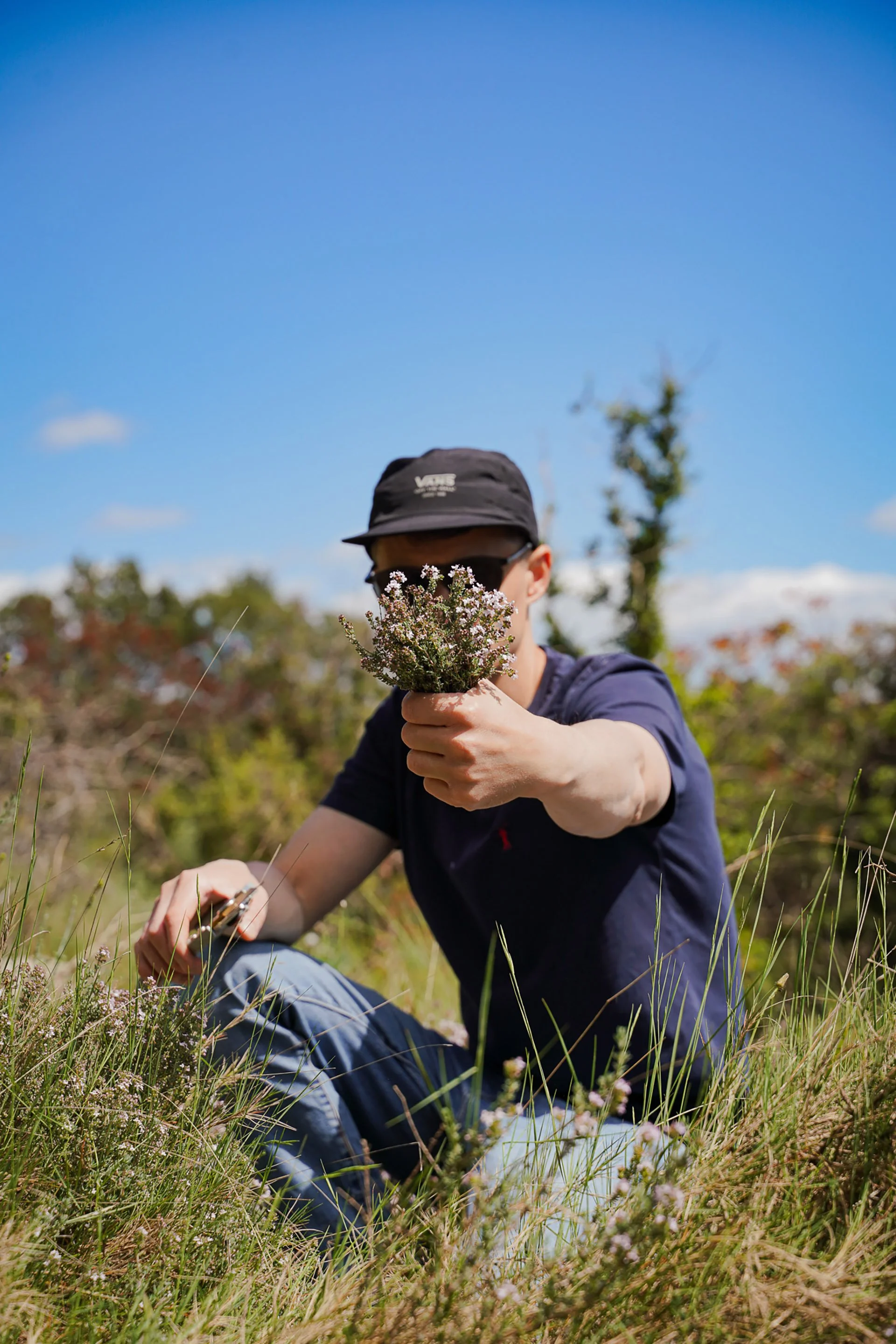 Chef Laurent Cherchi portant une casquette noire, des lunettes de soleil, et un t-shirt bleu, assise dans la garrigue, tenant un bouquet de fleurs de thym en main, sous un ciel bleu avec quelques nuages.