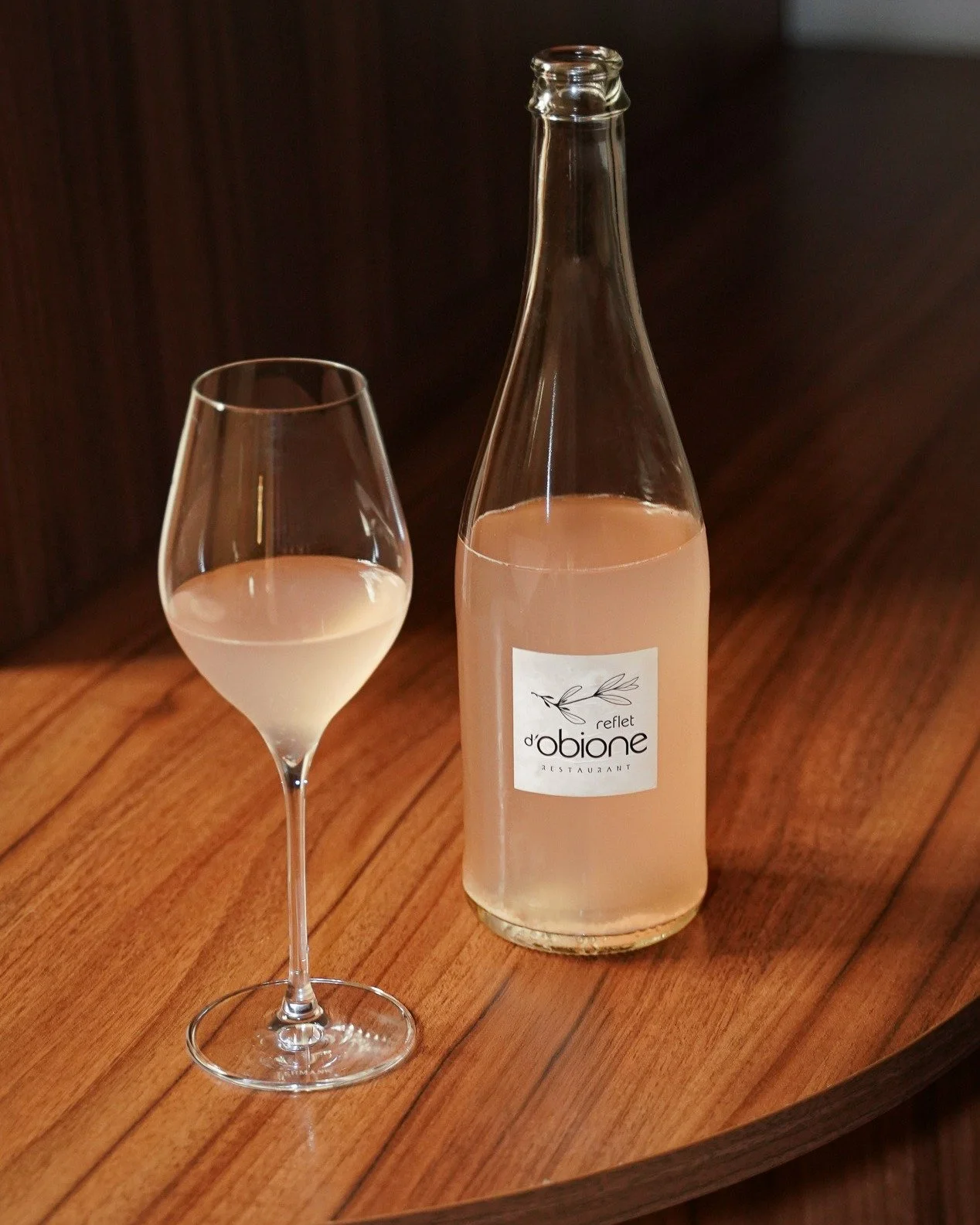 Une bouteille en verre avec une étiquette portant l'inscription 'reflet d'obione restaurant' et un verre à vin contenant un liquide rose clair, tous deux placés sur une surface en bois.
