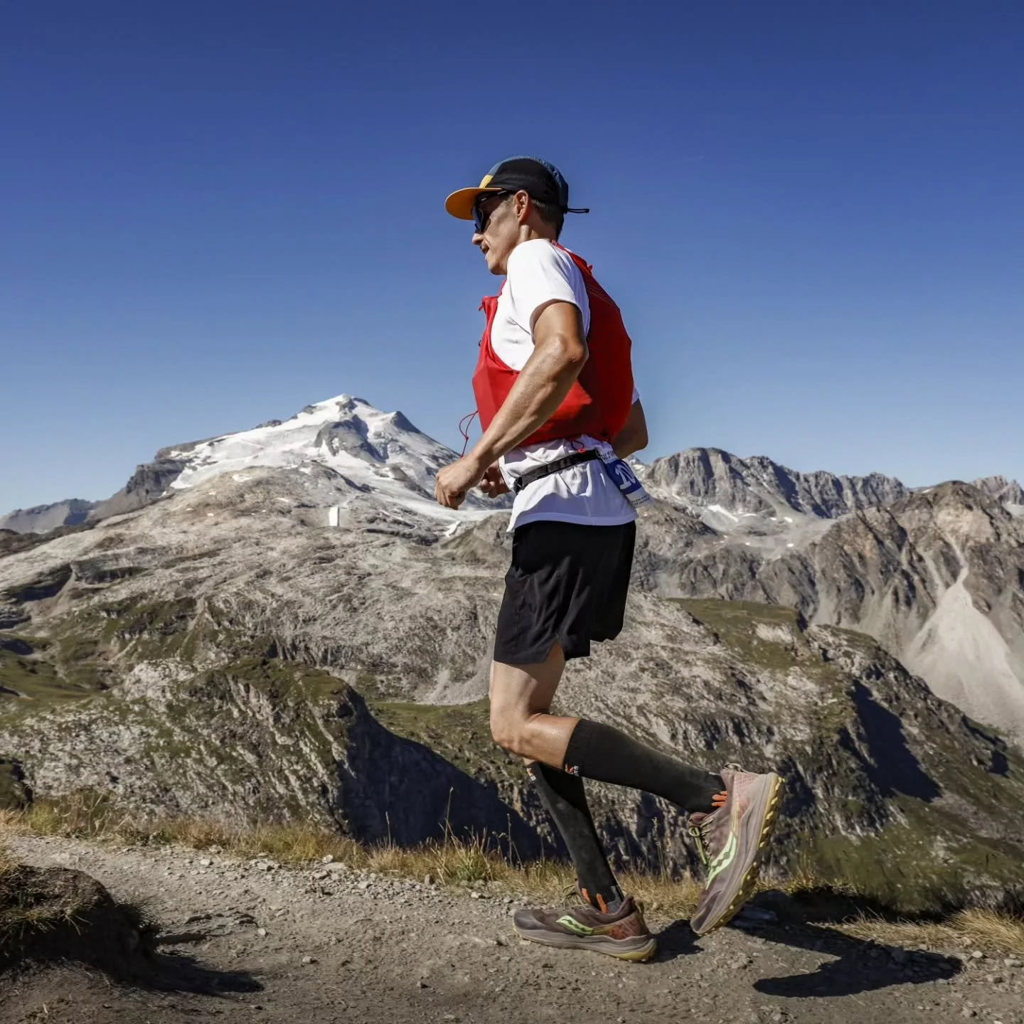 Chef Laurent Cherchi courant en montagne avec des montagnes enneigées en arrière-plan, portant une casquette, des lunettes de soleil, une veste rouge, un t-shirt blanc, un short noir, des chaussettes longues et des chaussures de course.