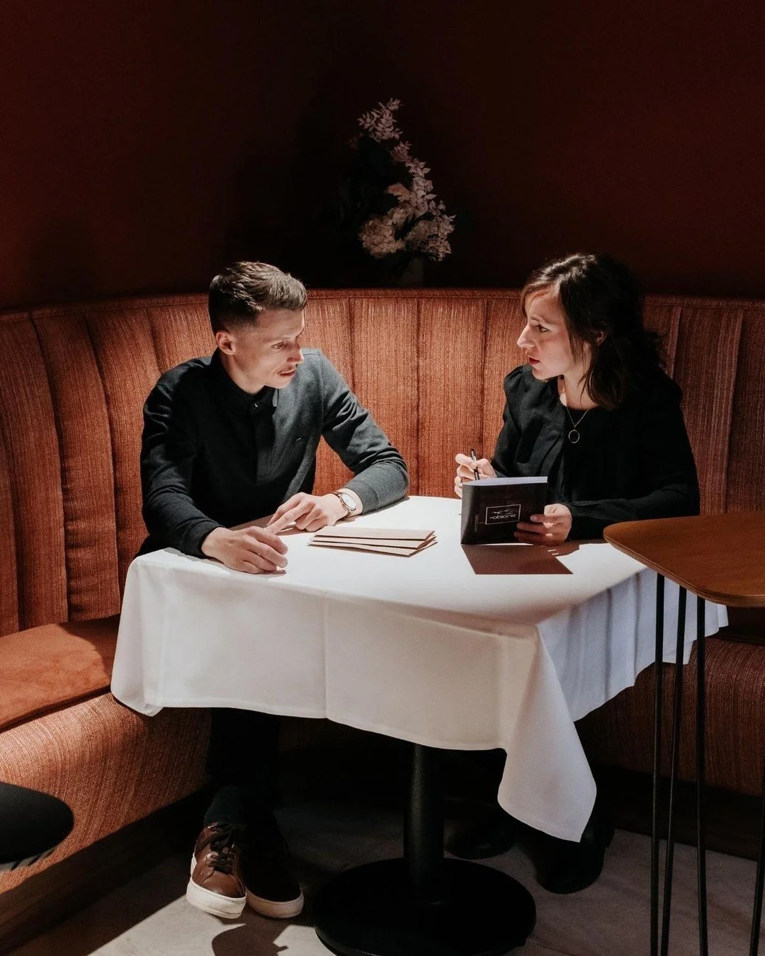 Chef Laurent Cherchi et Adèle Escafit directrice du restaurant Reflet d'Obione, assises à une petite table ronde avec une nappe blanche, avec un arrière-plan sombre et une décoration florale en arrière-plan.
