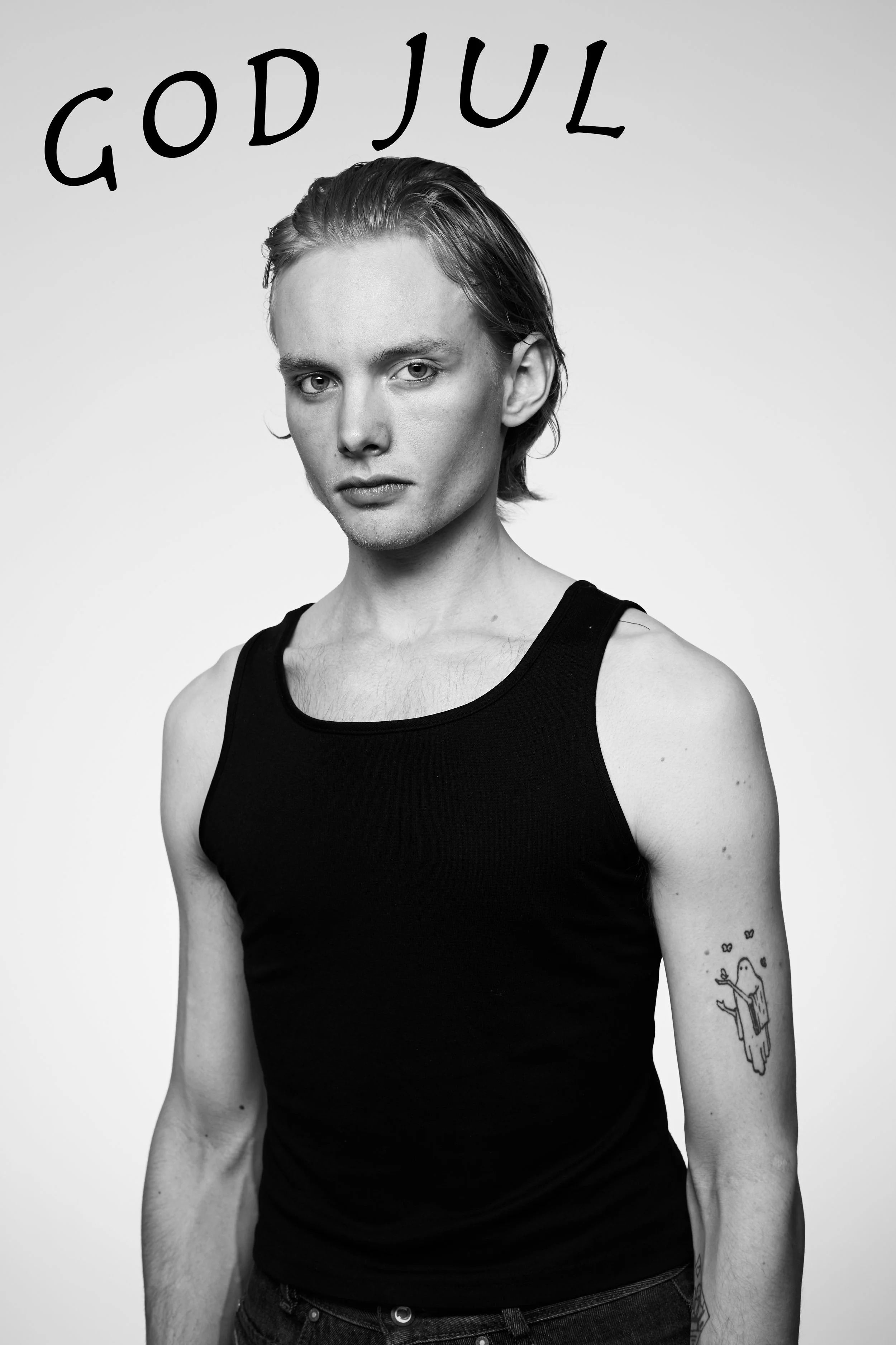 Black and white portrait of a young person with short hair wearing a sleeveless black shirt, with a tattoo of a ghost holding a vote on their left arm, and the words "GOD JUL" written in large font at the top.