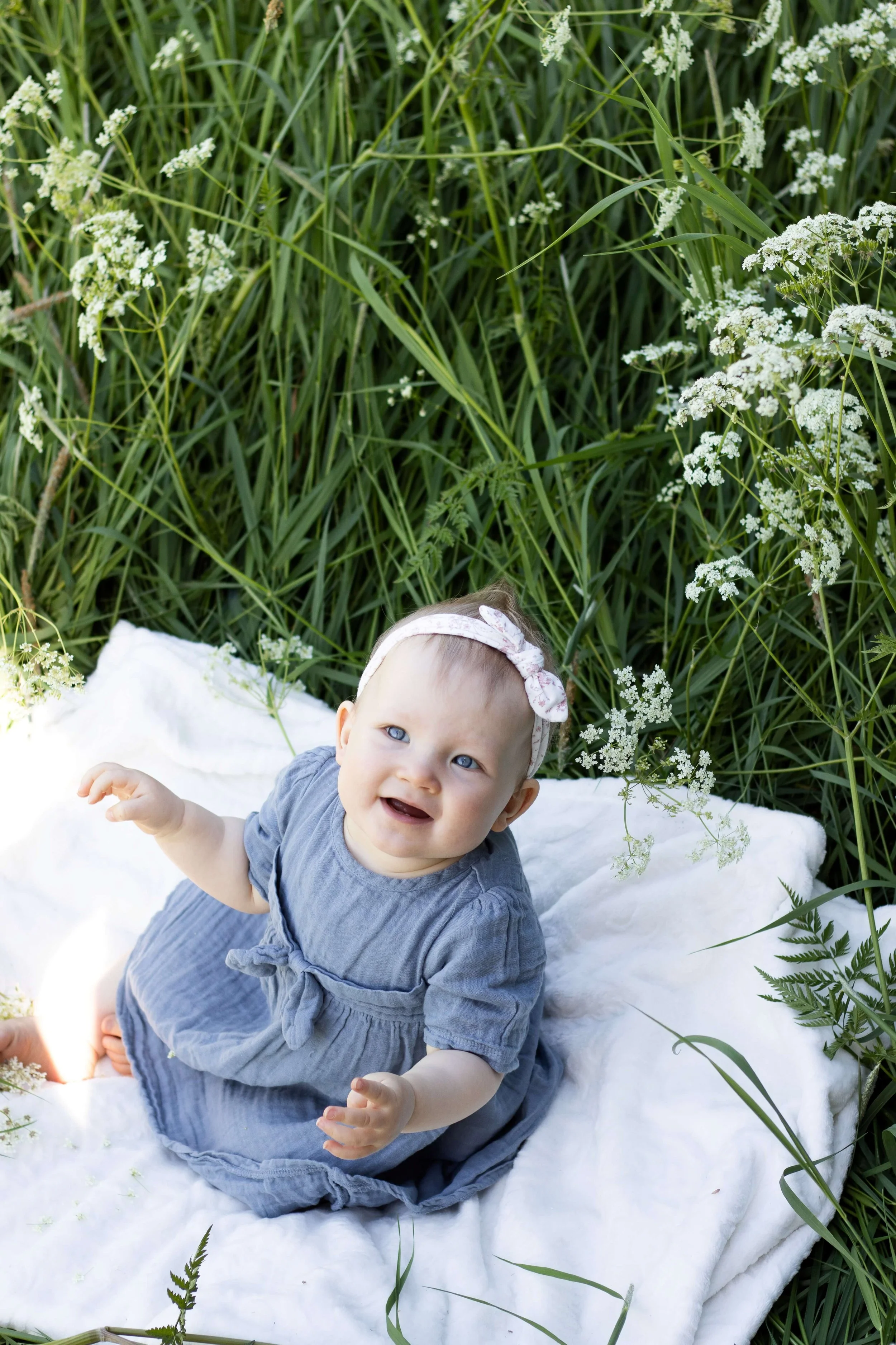 A smiling baby girl sitting on a white blanket outdoors among tall green grass and wildflowers, wearing a gray dress and a pink headband with a bow.