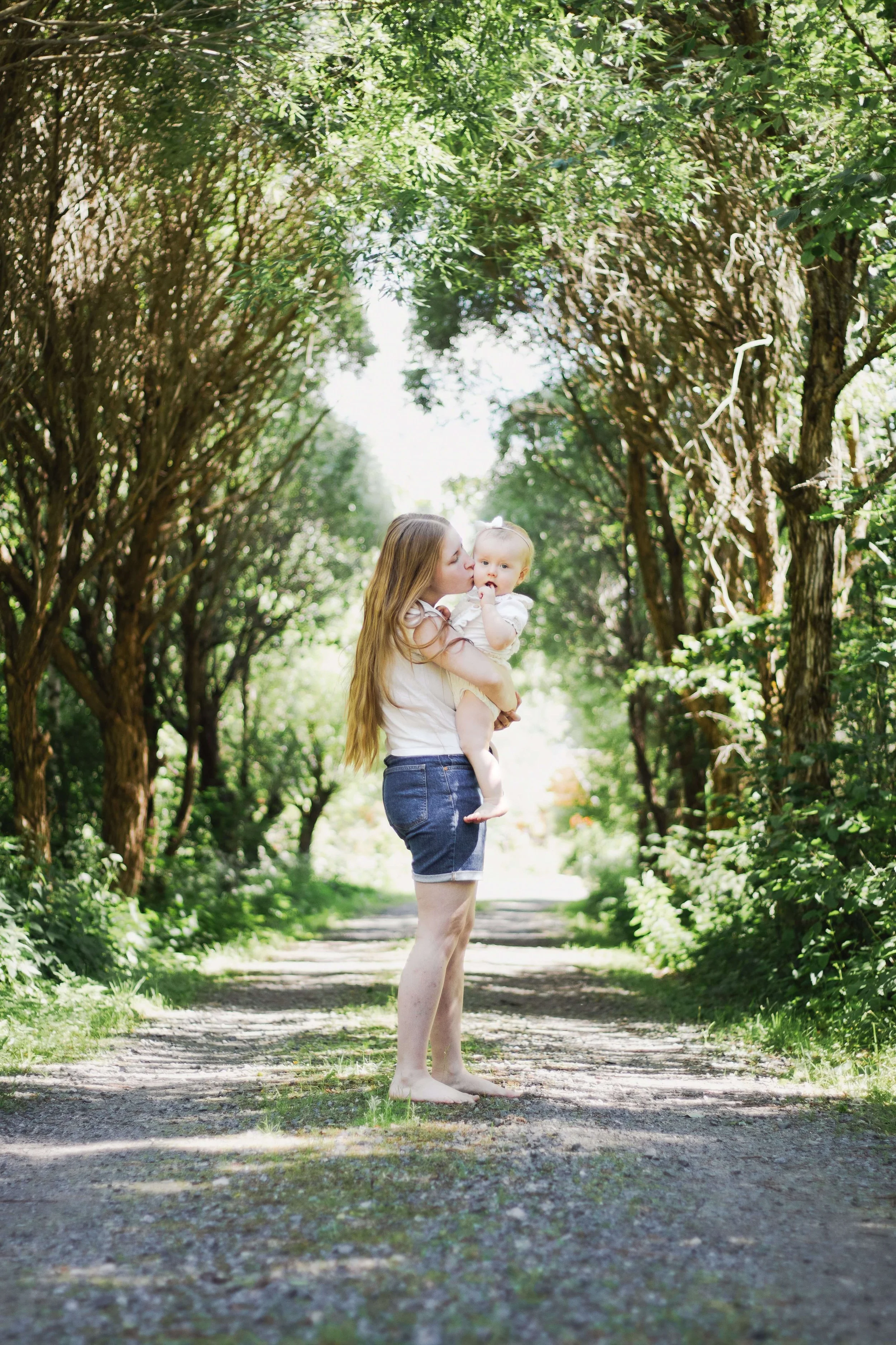 A woman with long hair, wearing a white shirt and denim shorts, stands barefoot on a forest path, holding a young child and kissing her on the cheek.