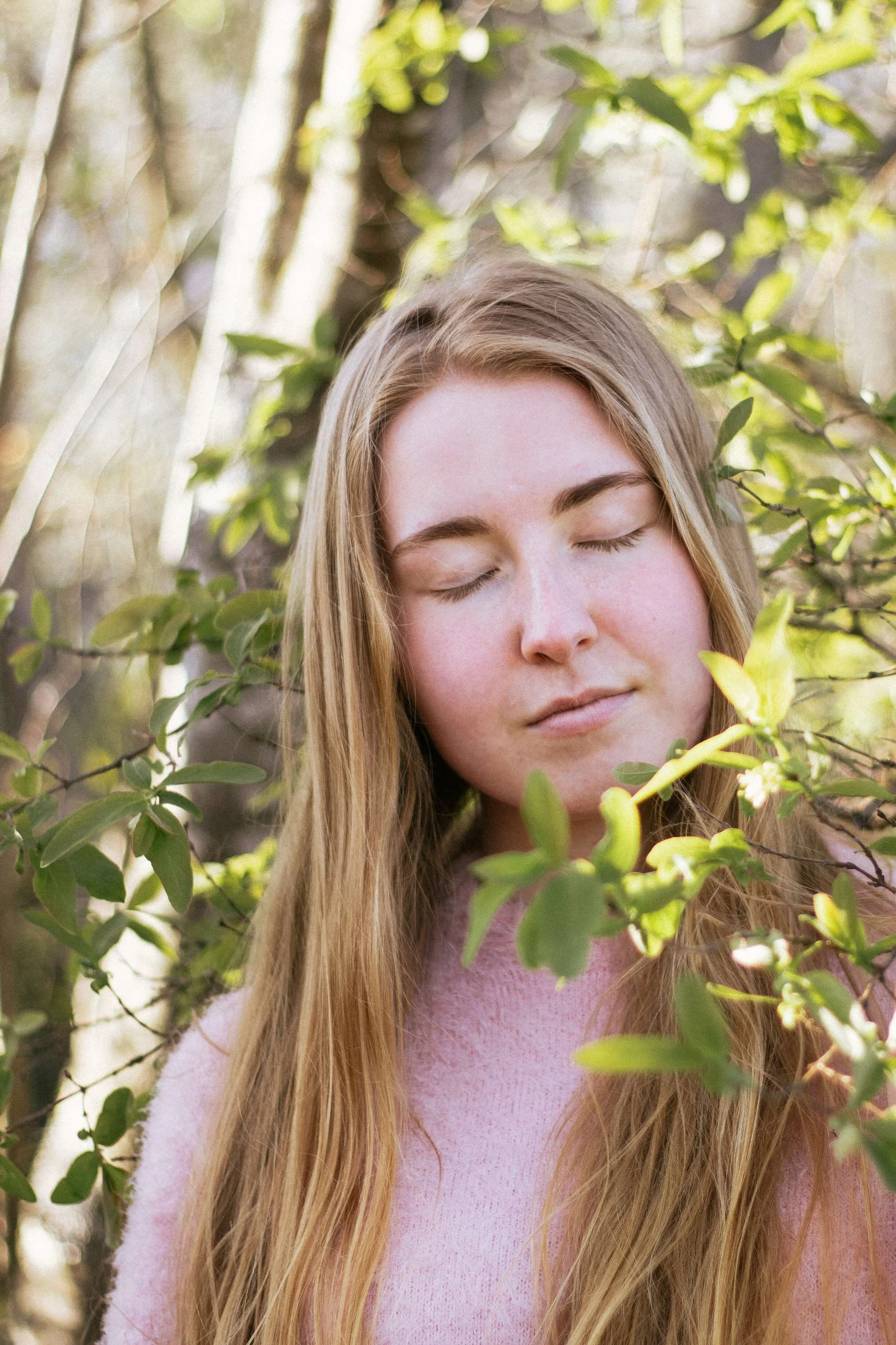 A young woman with long blonde hair and fair skin has her eyes closed and appears to be peacefully enjoying nature among green leafy branches in a sunny outdoor setting.