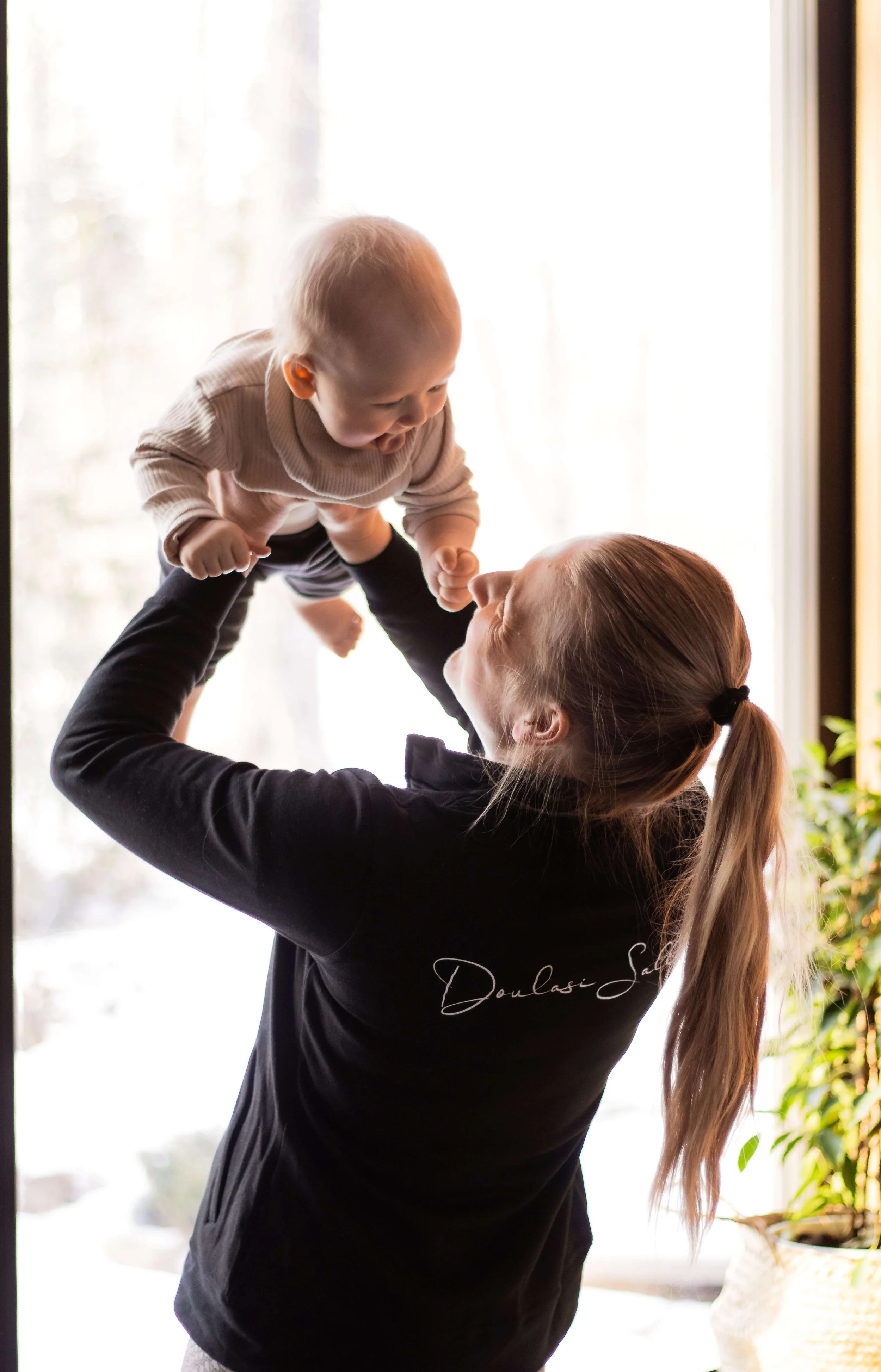 A woman lifting a baby in the air inside a room with a large window and a potted plant in the background. The woman has long hair in a ponytail and is wearing a black top, while the baby is dressed in a beige onesie.
