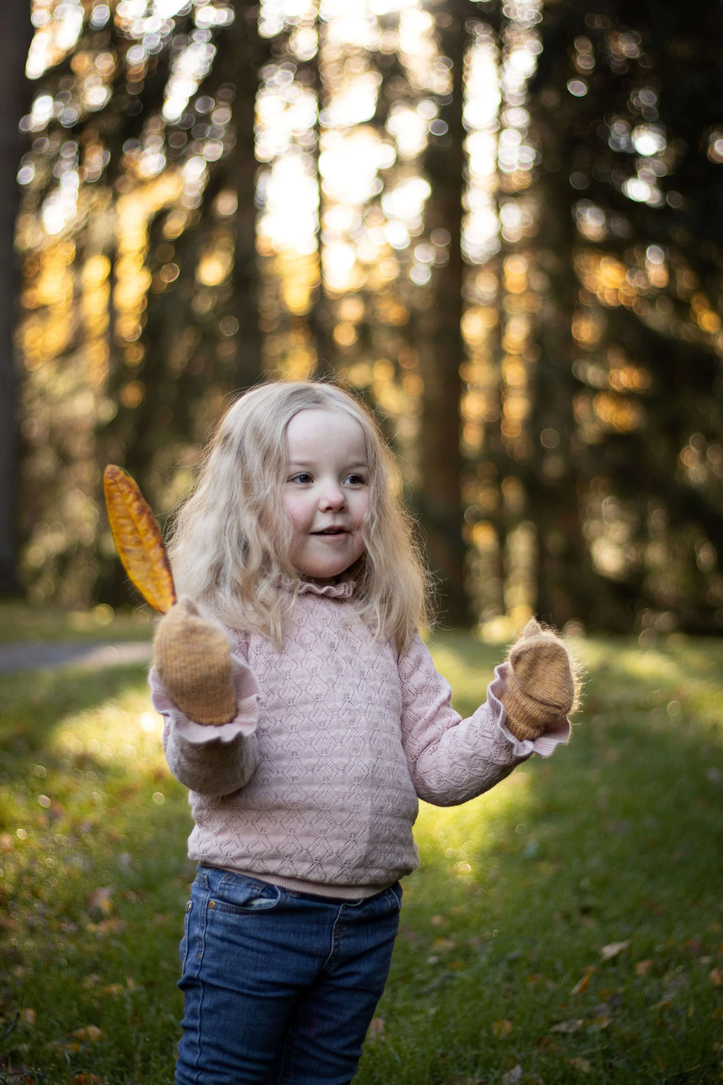 A young girl with blonde hair wearing a pink sweater, jeans, and brown gloves playing outdoors during autumn, holding a leaf in each hand, with trees and sunlight in the background.