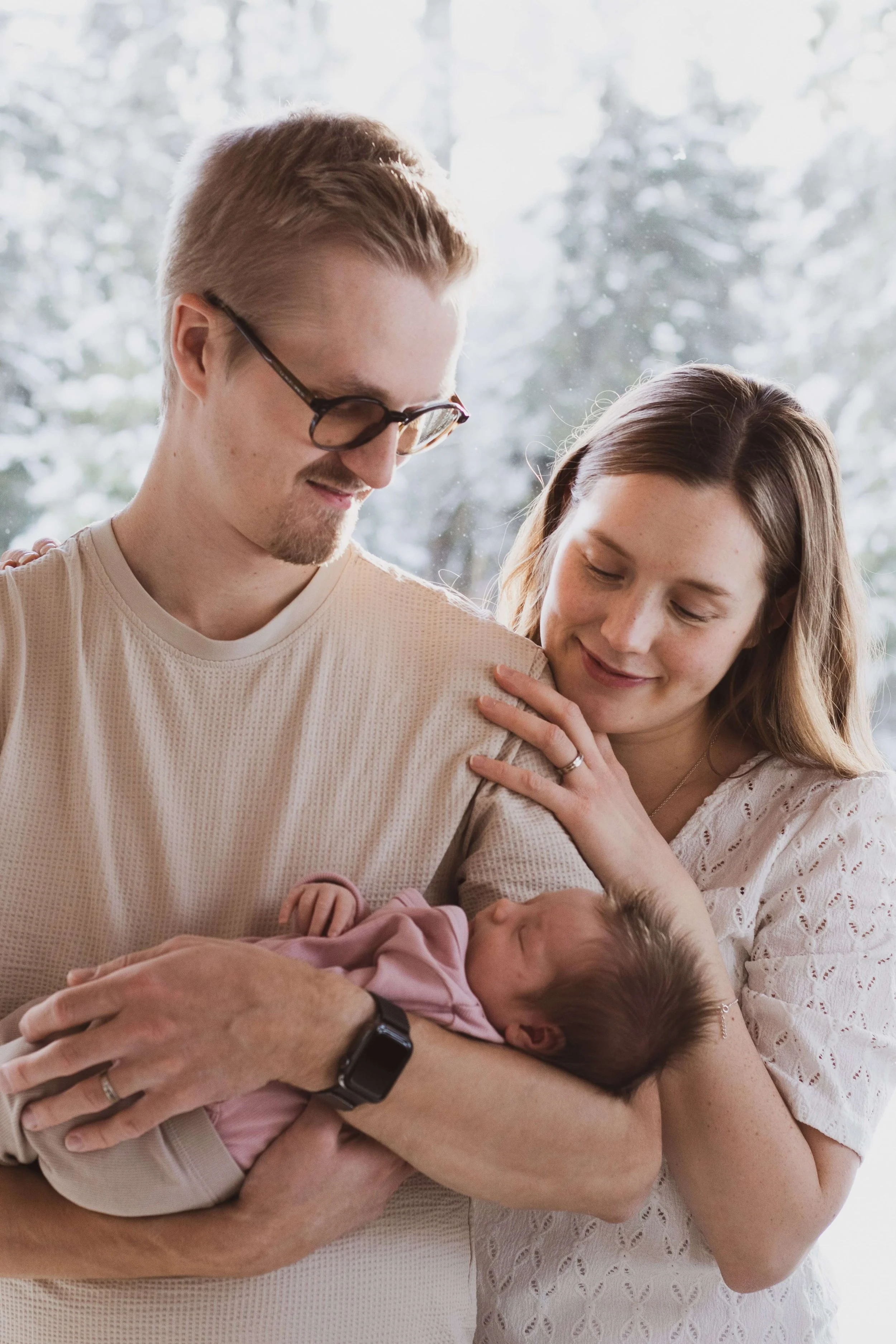 A couple holding a newborn baby, with warm, loving expressions, inside a bright room with a snowy outdoor scene visible through the window.