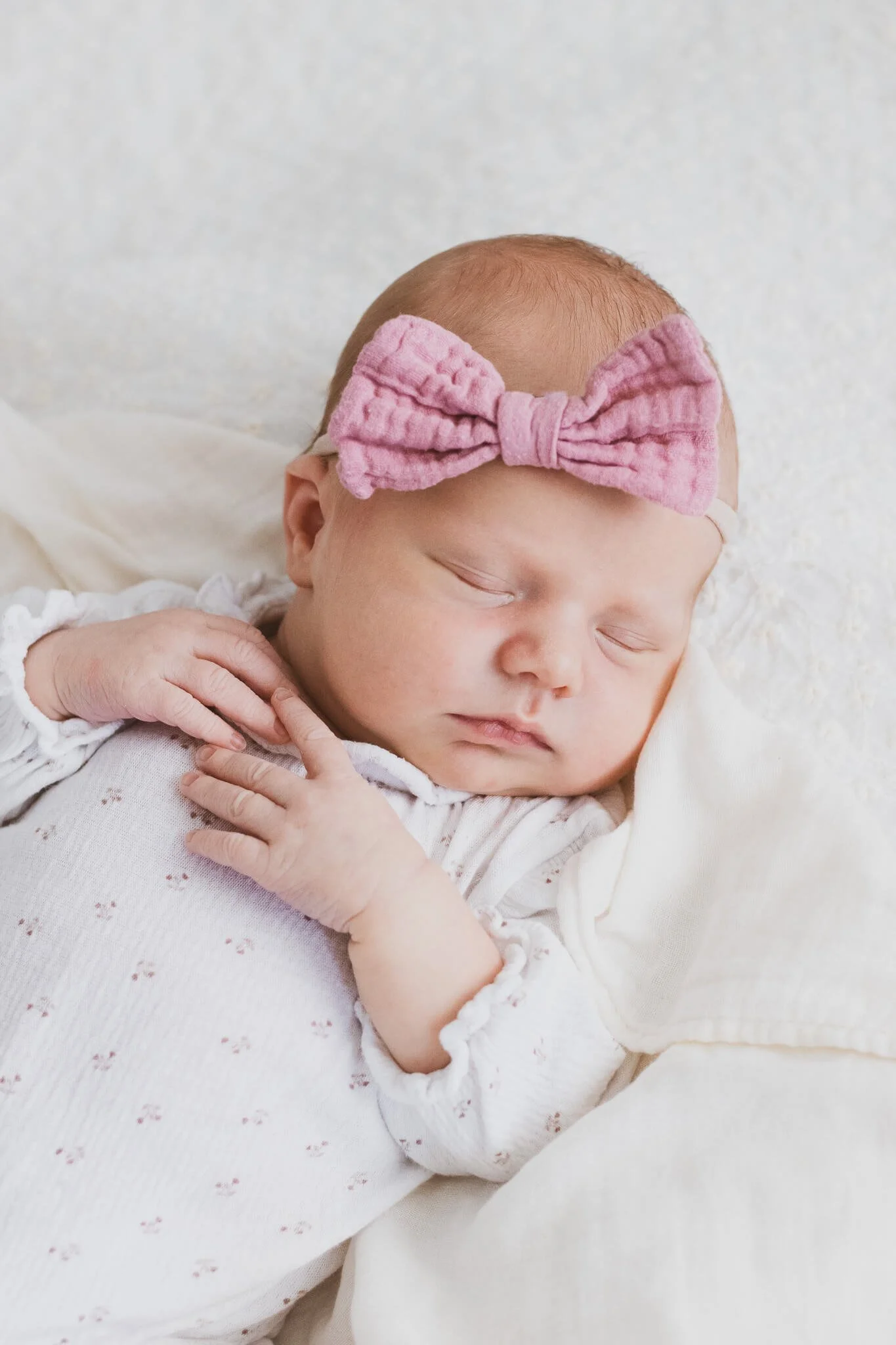 An adorable sleeping baby girl wearing a large pink bow headband, resting peacefully on a soft white blanket.