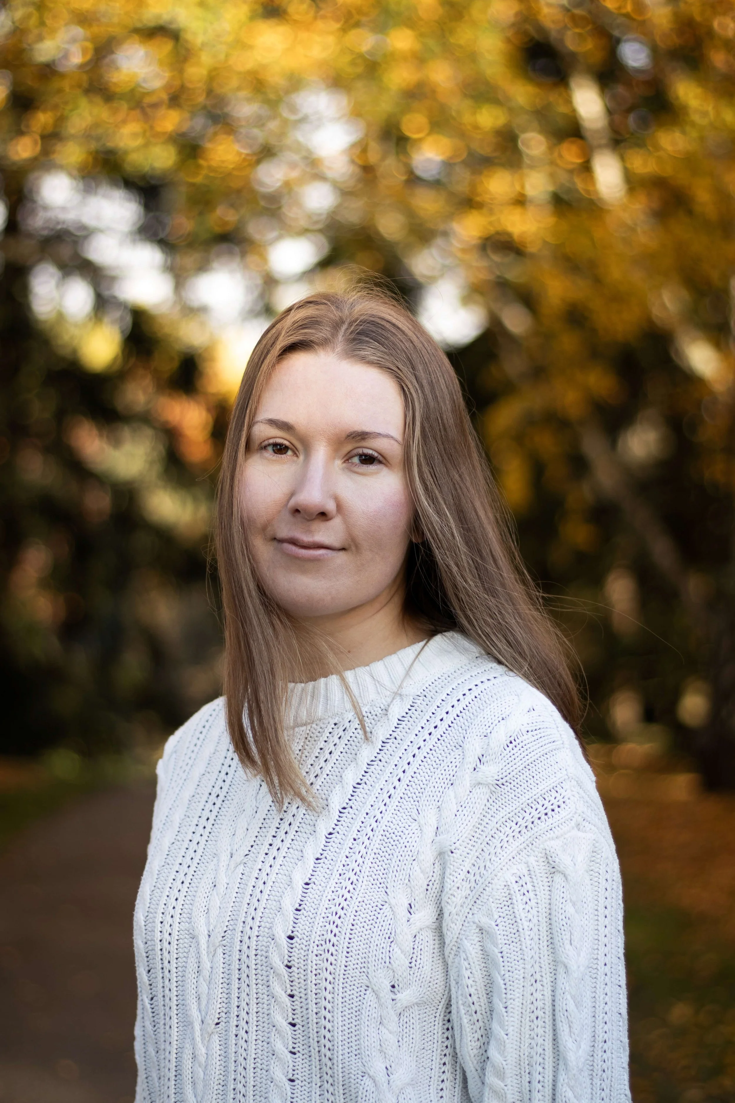 A woman with long light brown hair wearing a white knitted sweater outdoors during fall. She is standing on a path with blurred autumn trees and yellow leaves in the background.