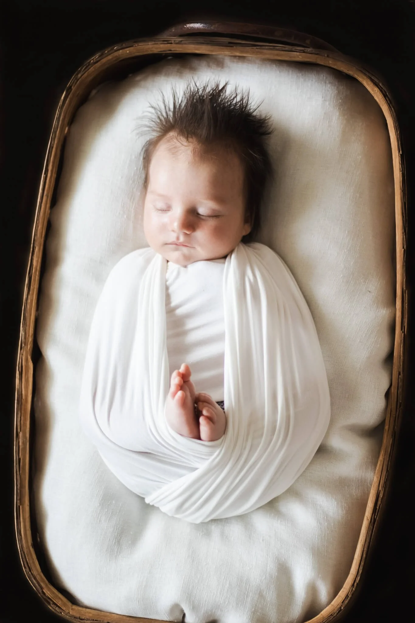 A sleeping baby wrapped in white swaddling cloth, lying on a cushion in a wooden bassinet.