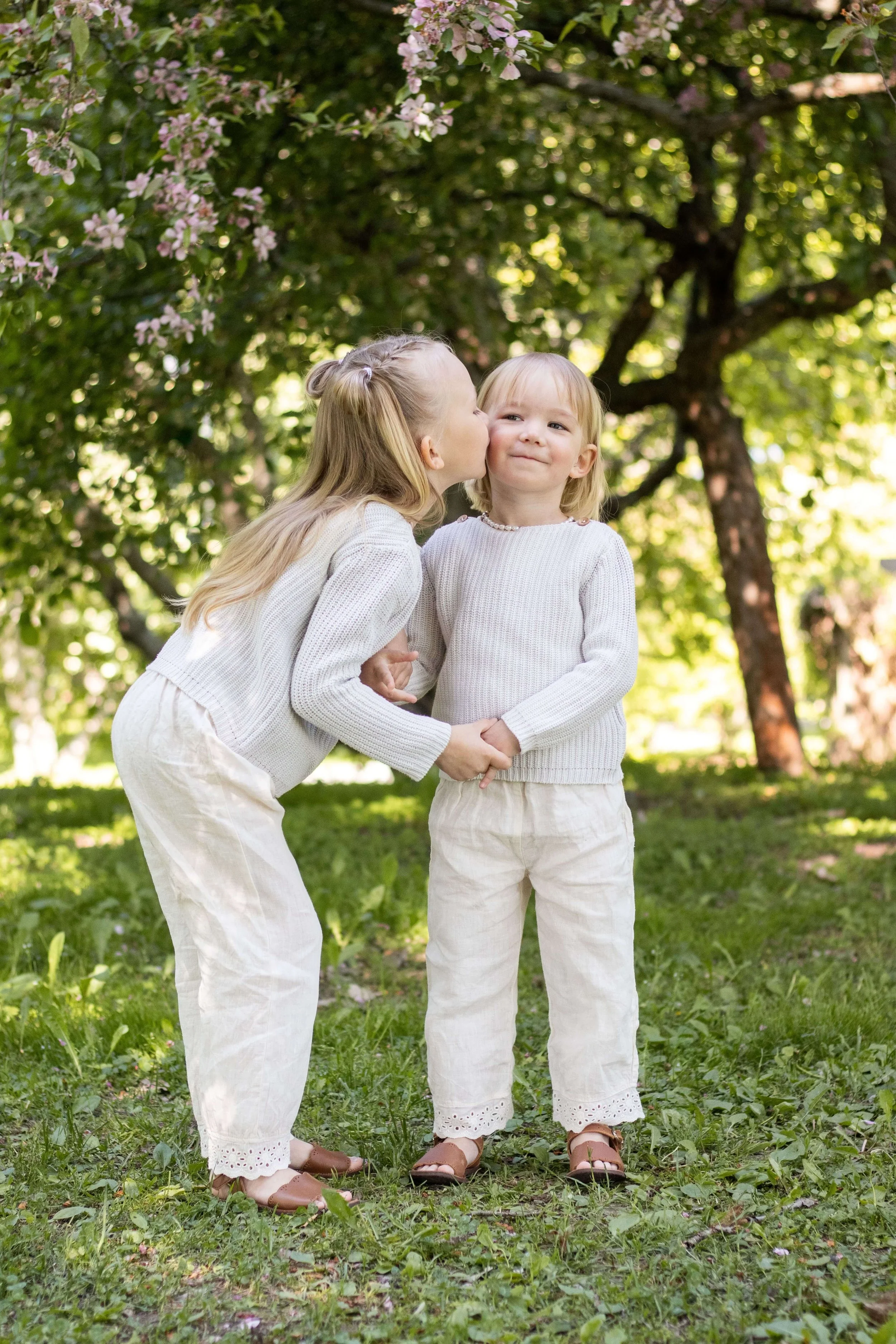 Two young girls, one kissing the other on the cheek, standing outdoors in a grassy area with trees and pink blossoms in the background, dressed in light-colored clothing.