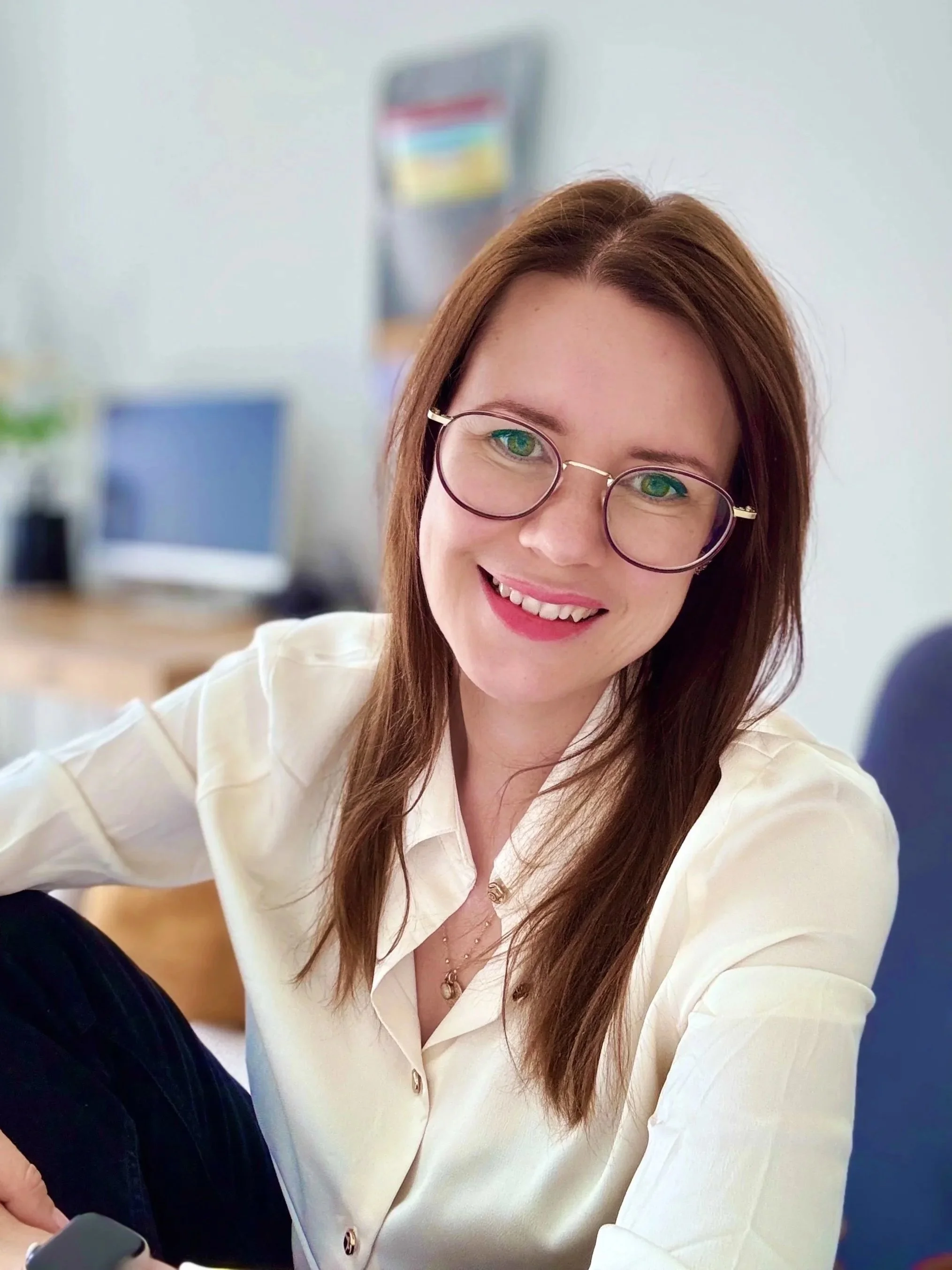 Femme souriante portant des lunettes rondes, vêtue d'une chemise blanche, assise dans un bureau moderne.