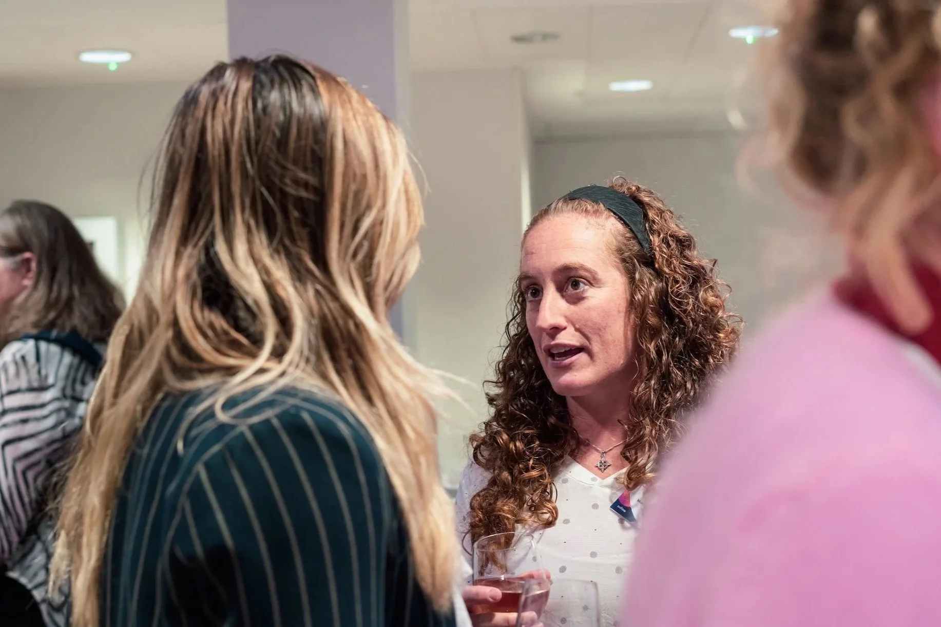 Three women engaged in a conversation, one in focus holding a glass of wine, woman in the middle with long curly hair and a headband, indoor setting with ceiling lights.