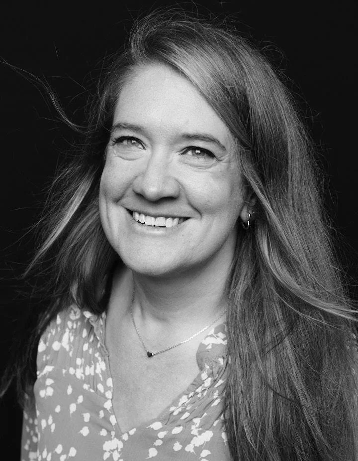 Black and white portrait of a smiling woman with long hair, wearing a patterned blouse, earrings, and a necklace, against a dark background.