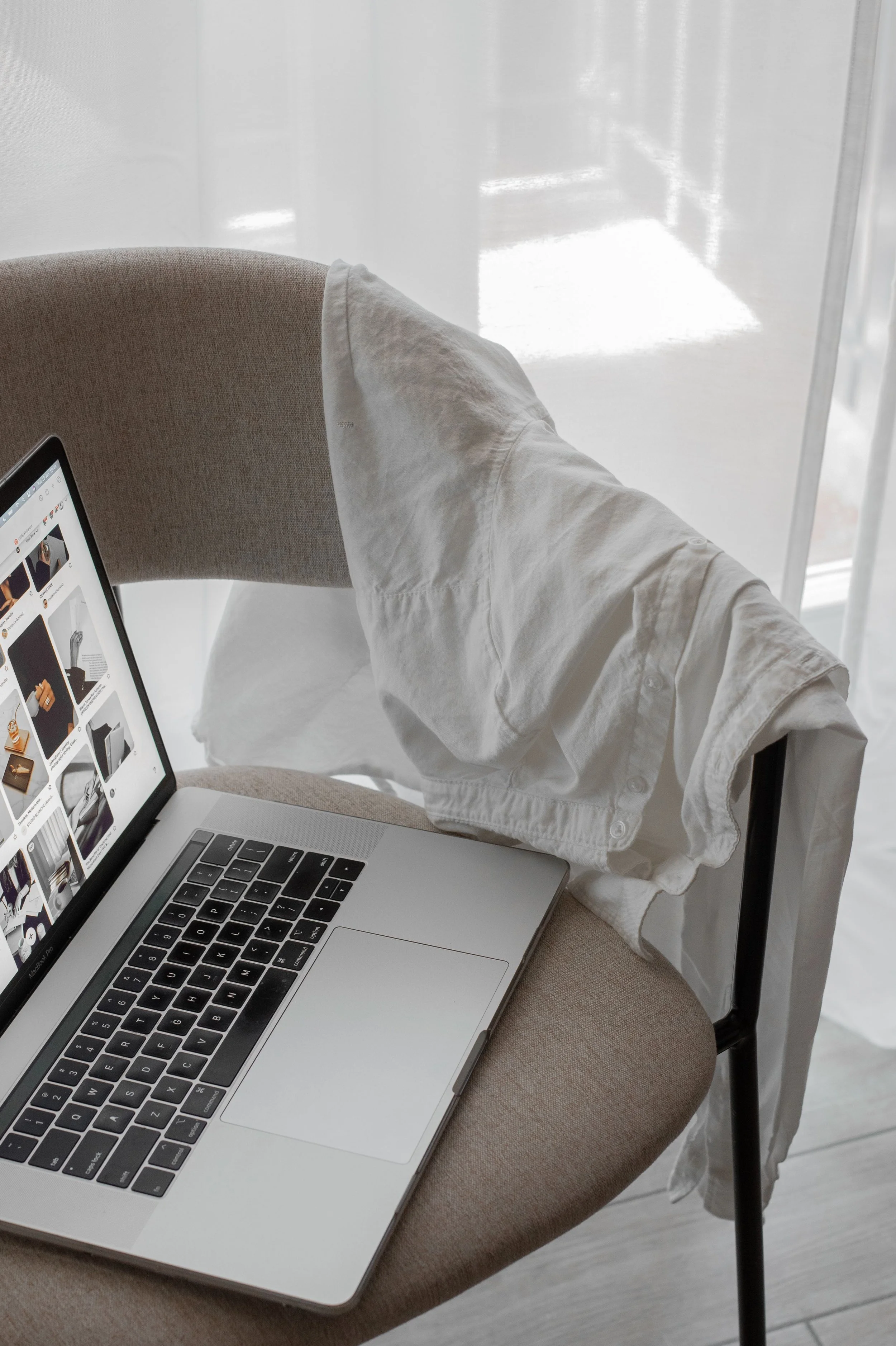 A laptop on a beige chair with a white shirt draped over the back, near a window with white curtains.