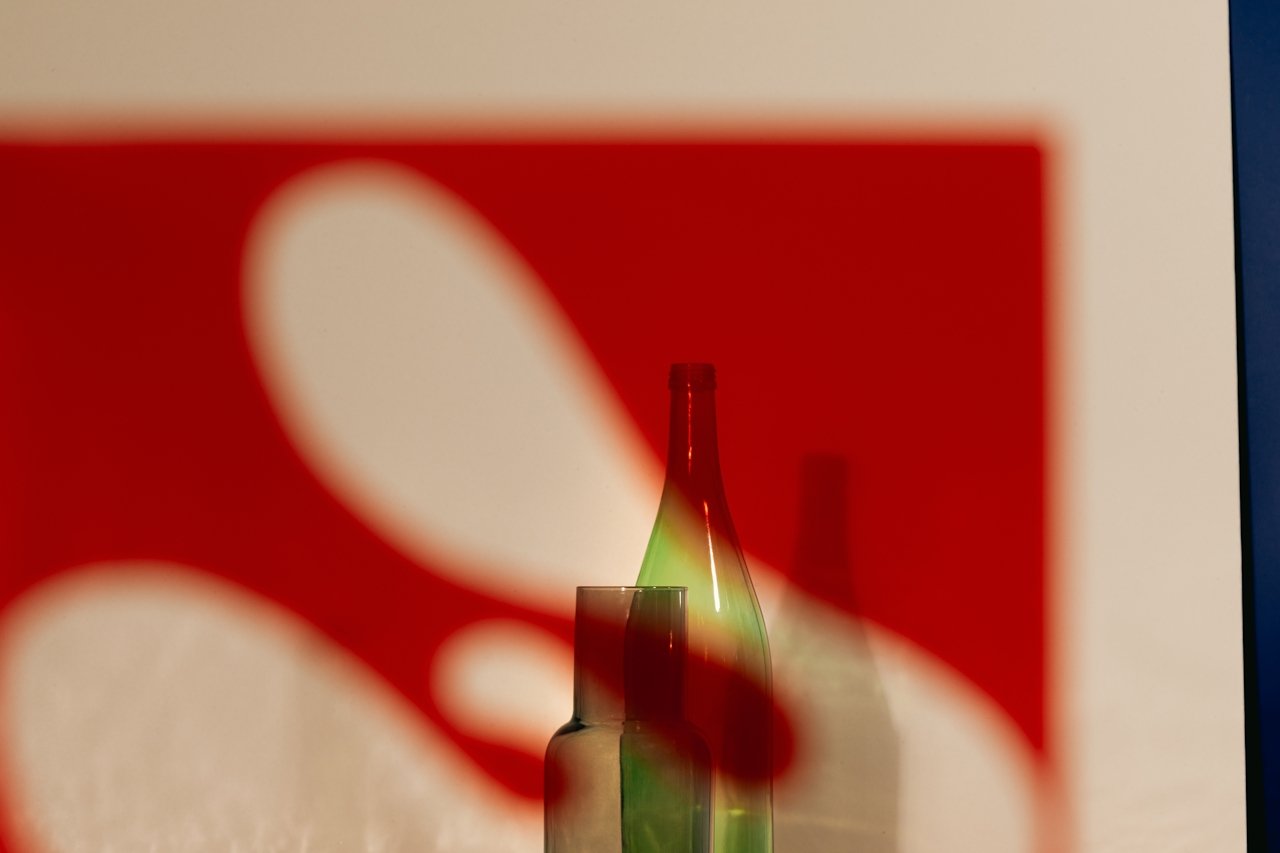 Close-up of a green glass bottle behind a red card with white abstract shapes, casting a shadow on the card.