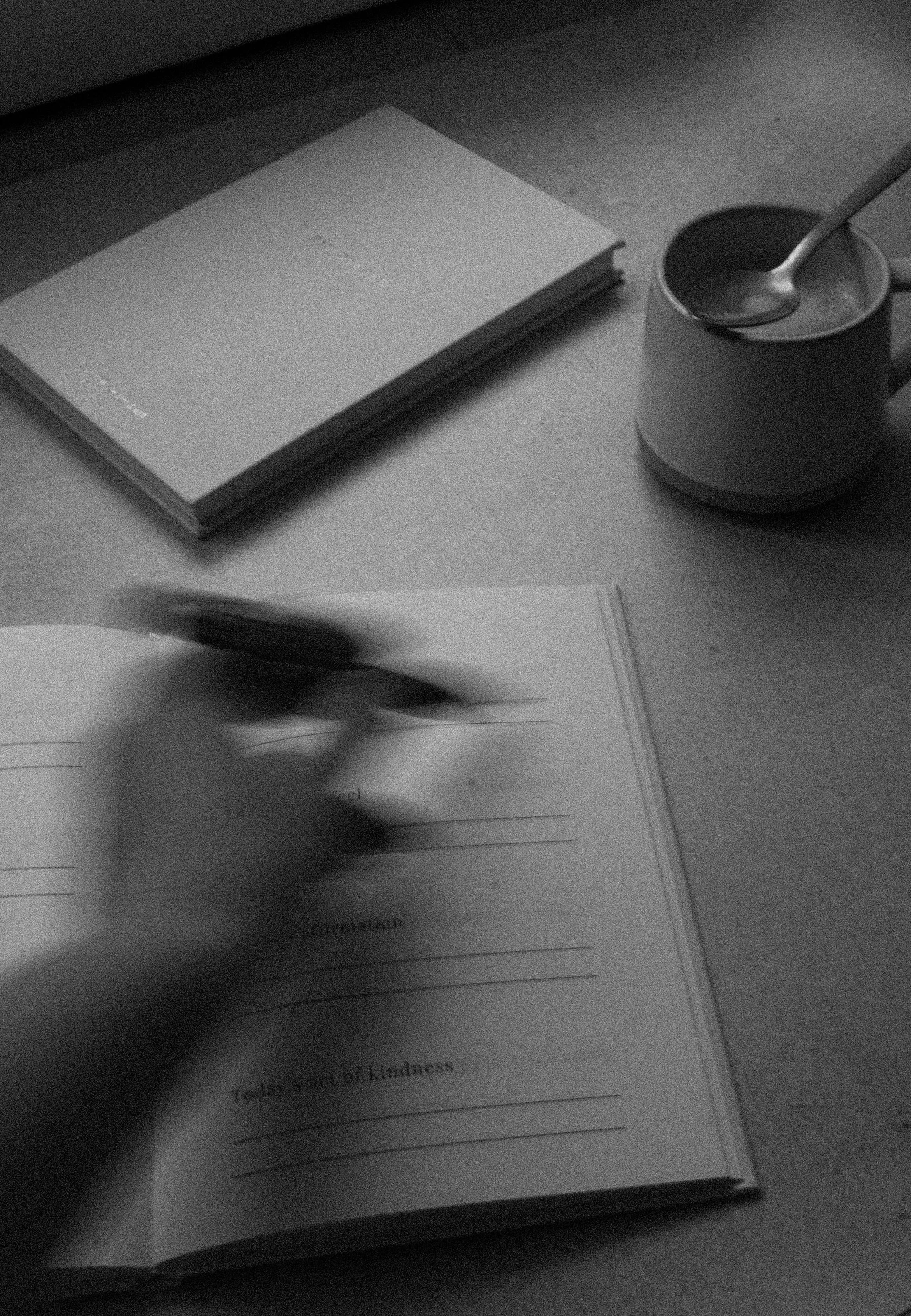 A person writing in a notebook at a desk with a stack of notebooks, a cup of tea or coffee with a spoon, and an open book.