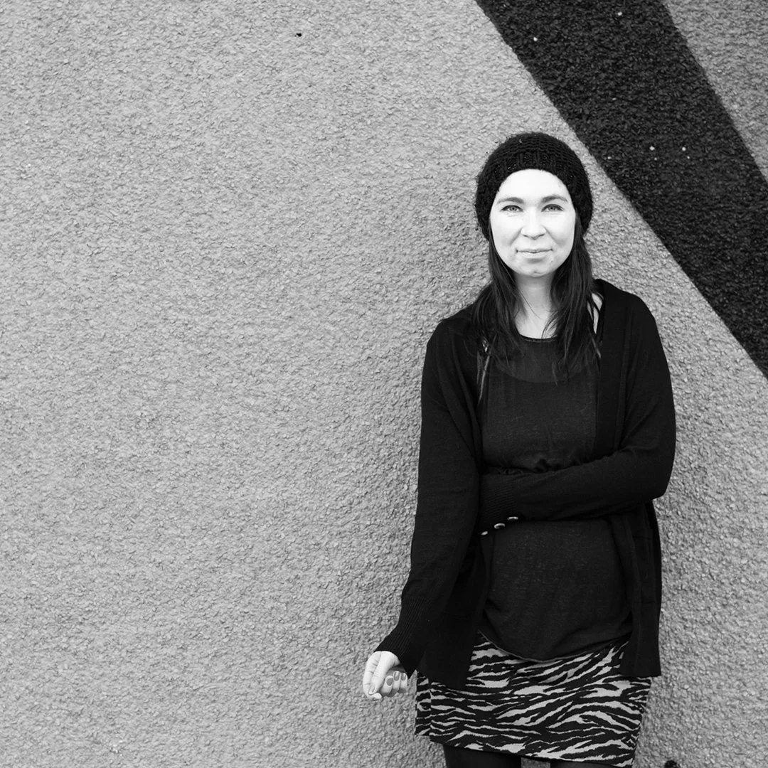 Black and white photo of a woman standing with arms crossed in front of a textured wall with a dark diagonal stripe.