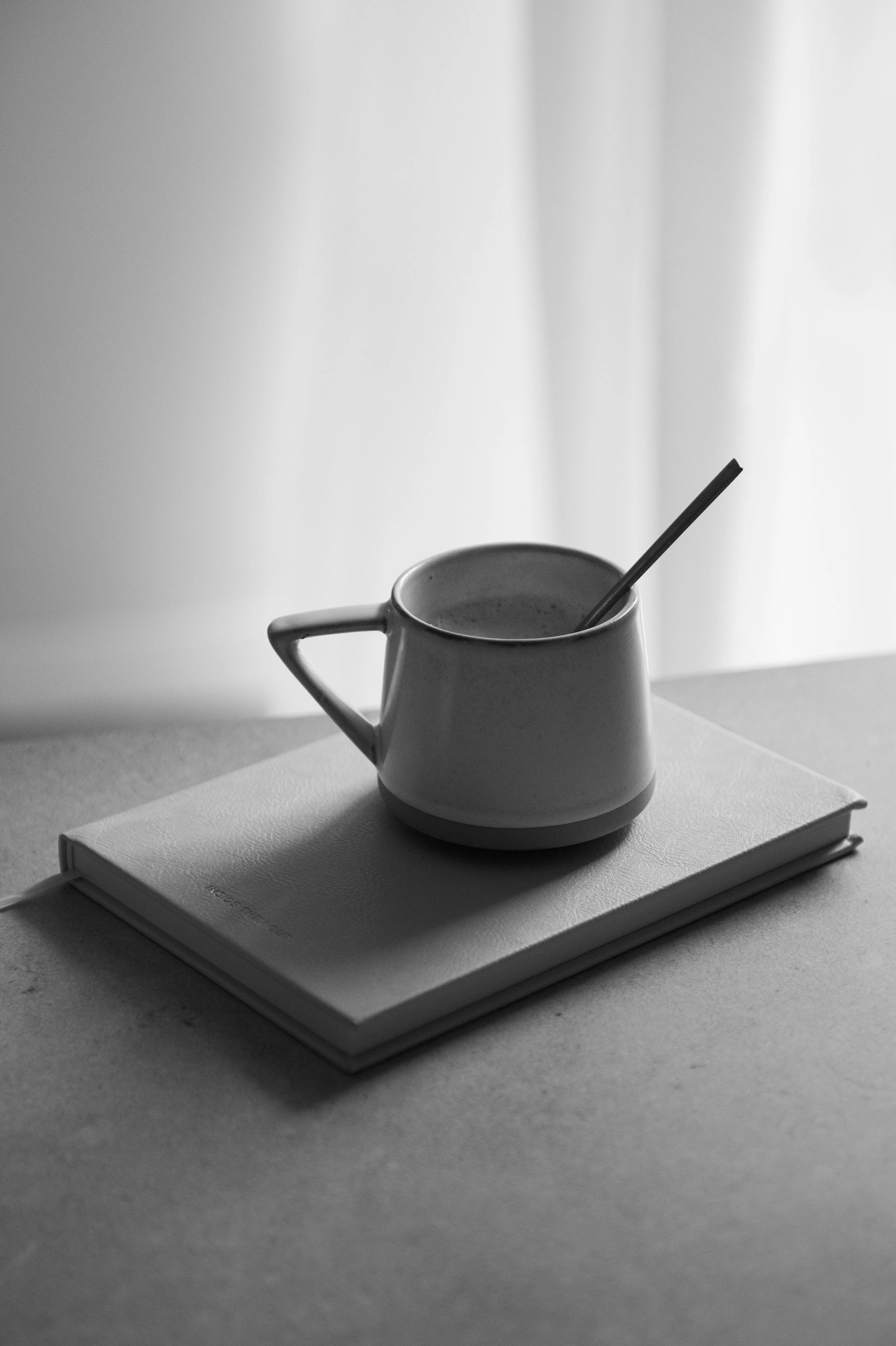 A black-and-white photo of a mug with a spoon in it, placed on top of a closed notebook on a table, with a blurred curtain in the background.